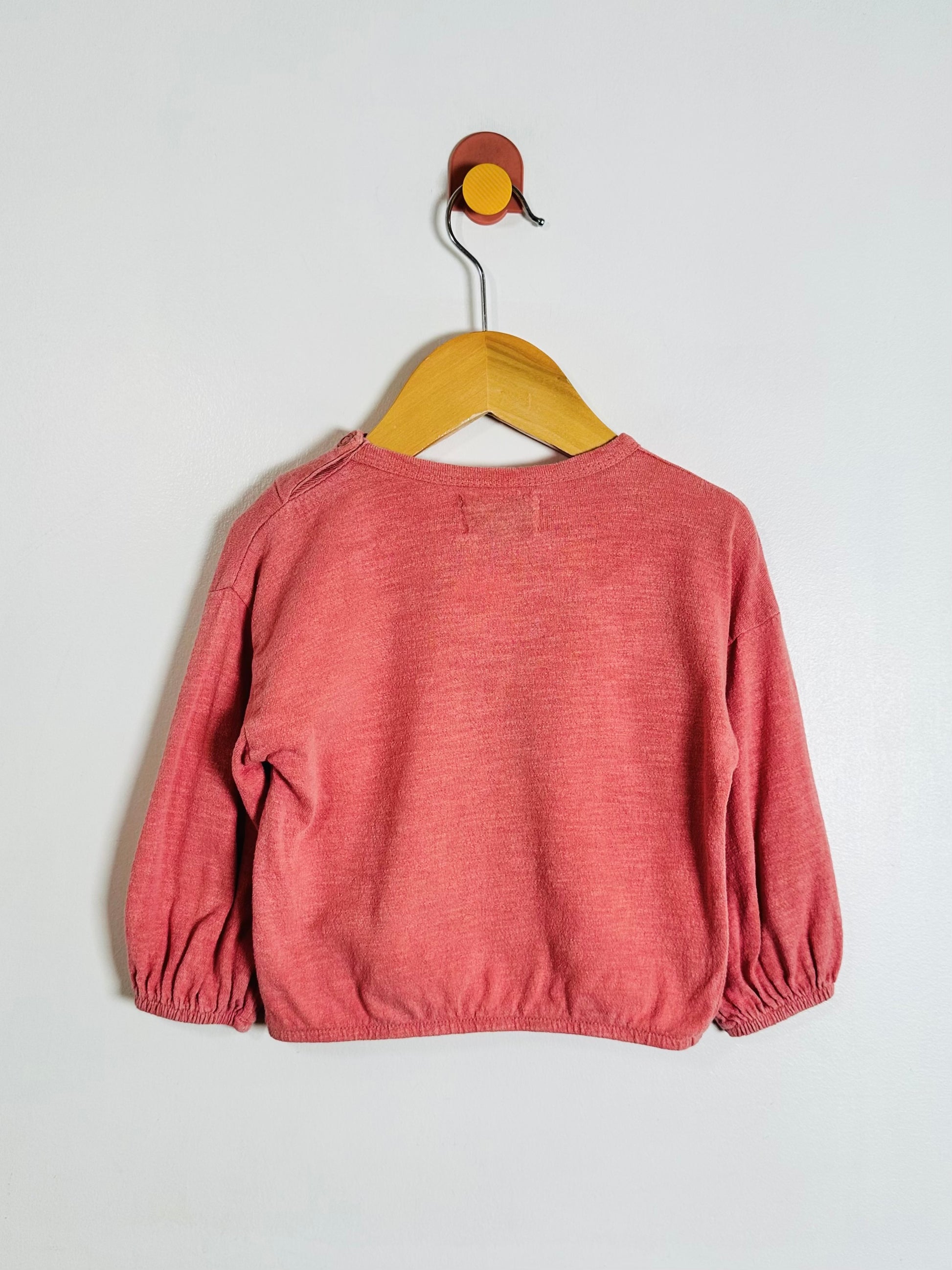 A dusty rose long-sleeve knit top for toddlers, hanging on a wooden hanger from a wall hook against a plain light gray wall.