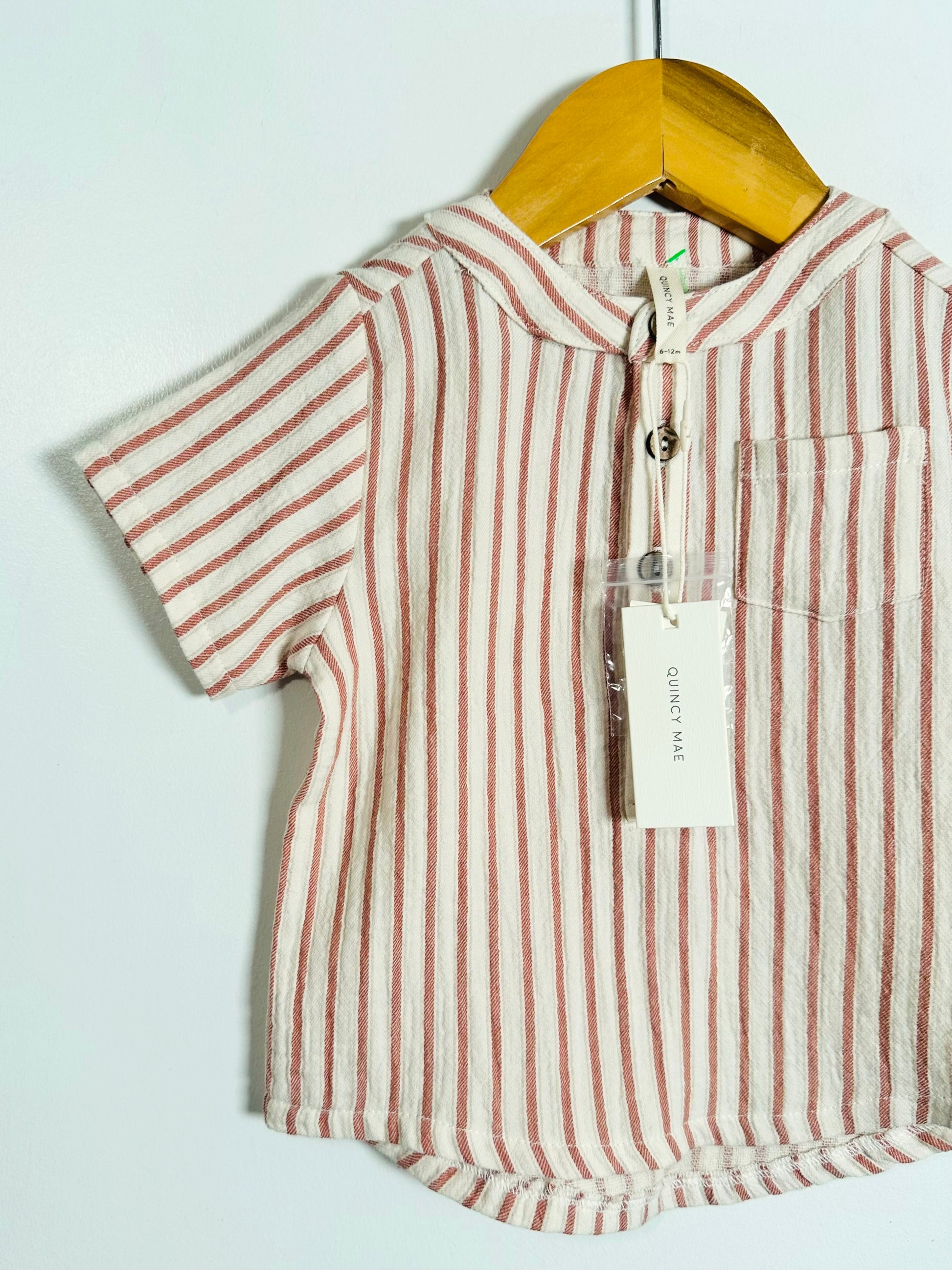 A short-sleeve striped polo shirt for kids, featuring vertical stripes in dusty pink and cream, hanging on a wooden hanger against a plain white wall. The shirt has a buttoned collar, a chest pocket, and a paper brand tag that reads 'QUINTY MALL'.