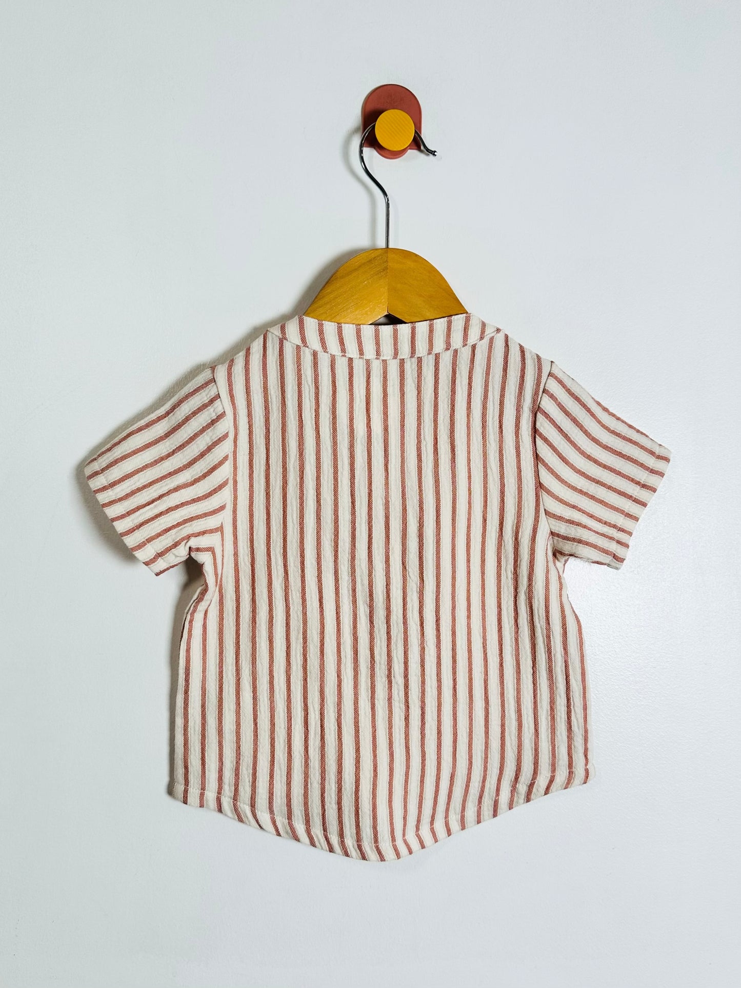 A short-sleeve baby shirt with vertical brown stripes on a cream linen-like fabric, hanging on a wooden hanger against a light gray wall.