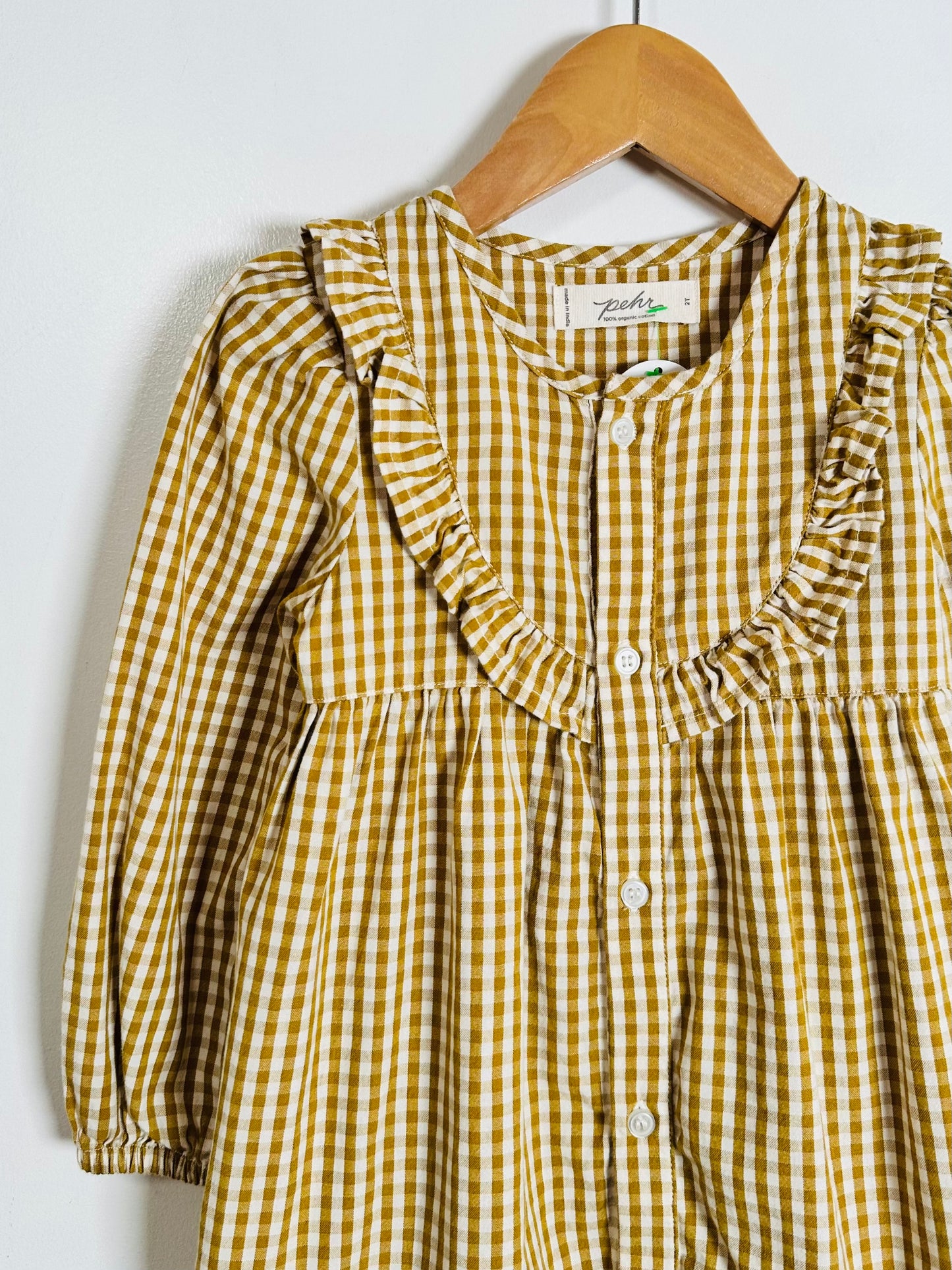 A yellow and white gingham long-sleeve blouse with ruffled trim, hanging on a wooden hanger against a plain white wall.