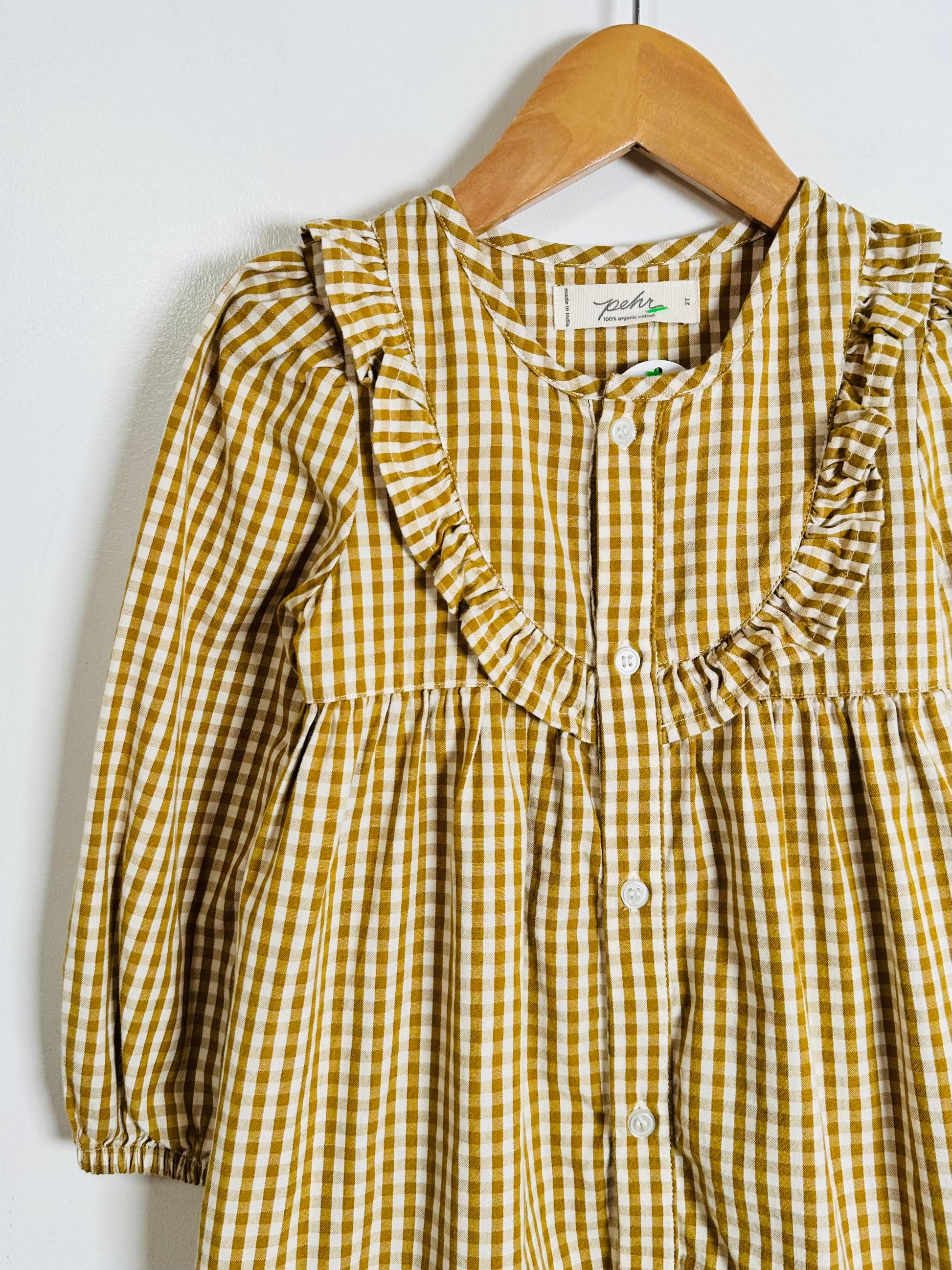 A yellow and white gingham long-sleeve blouse with ruffled trim, hanging on a wooden hanger against a plain white wall.