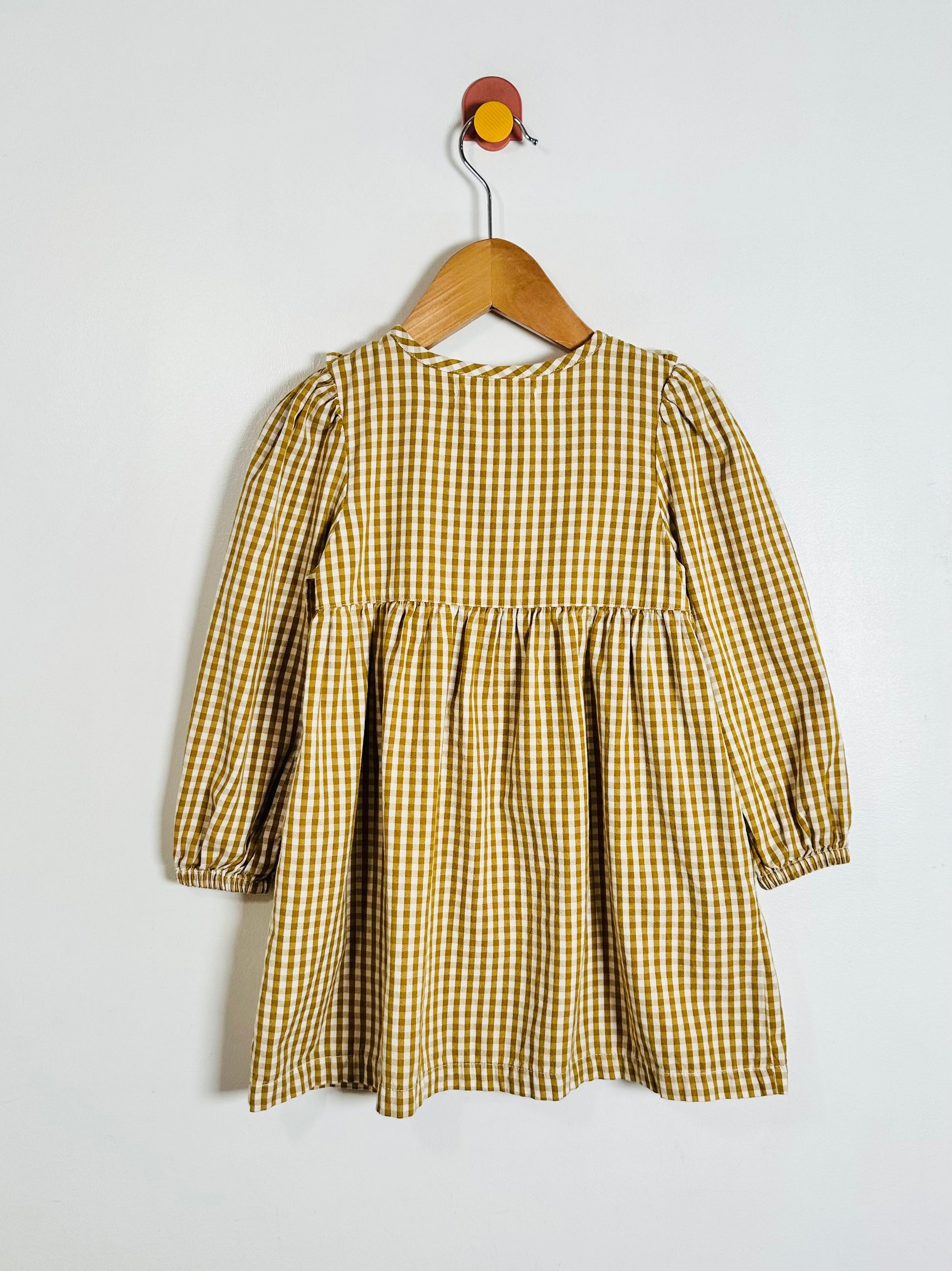 A toddler's long-sleeve dress with a yellow and white gingham check pattern, hanging on a wooden hanger against a plain light gray wall.