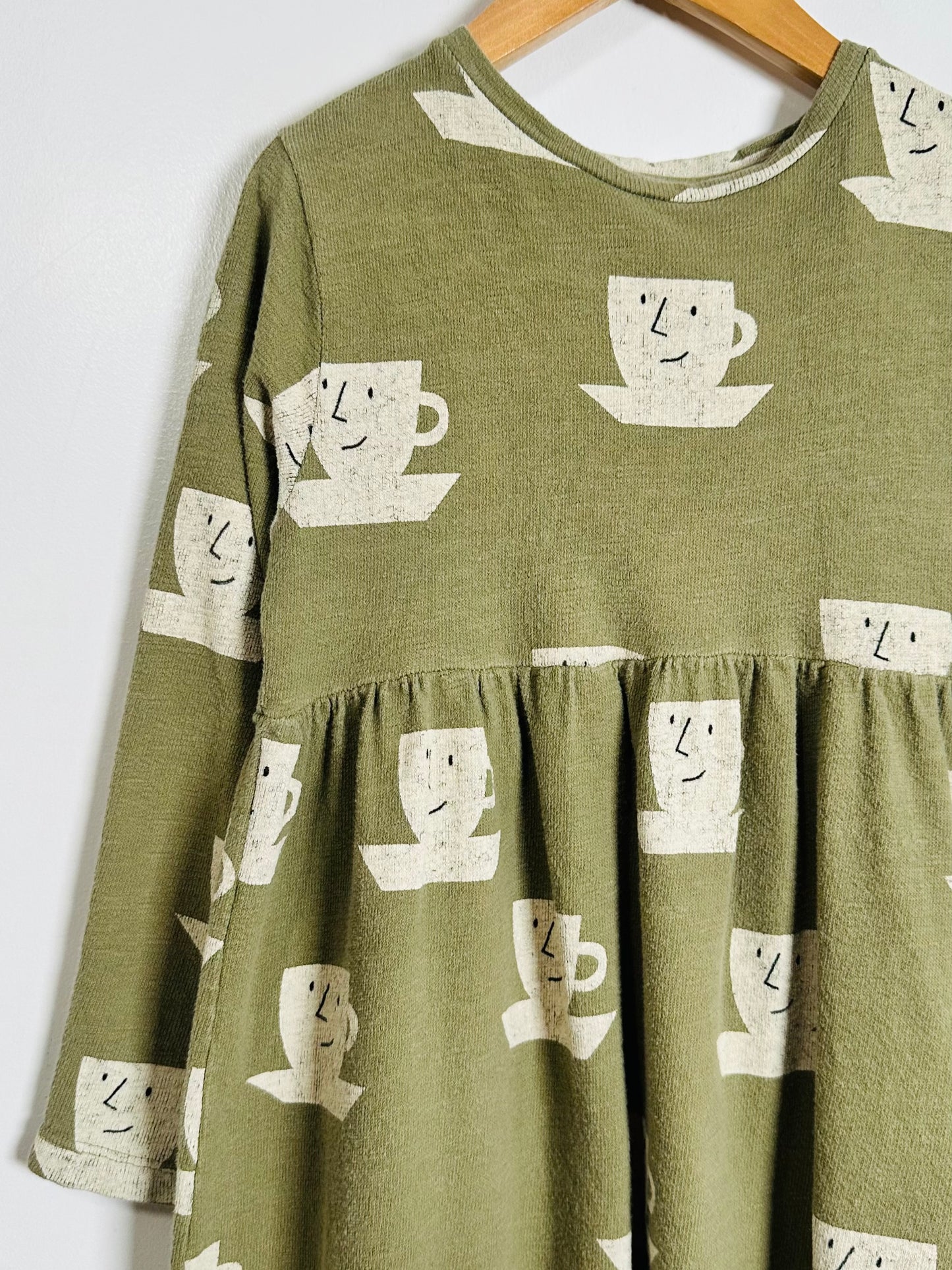 An olive green tiered long-sleeve dress with a white coffee cup pattern, hanging on a wooden hanger against a plain white wall.