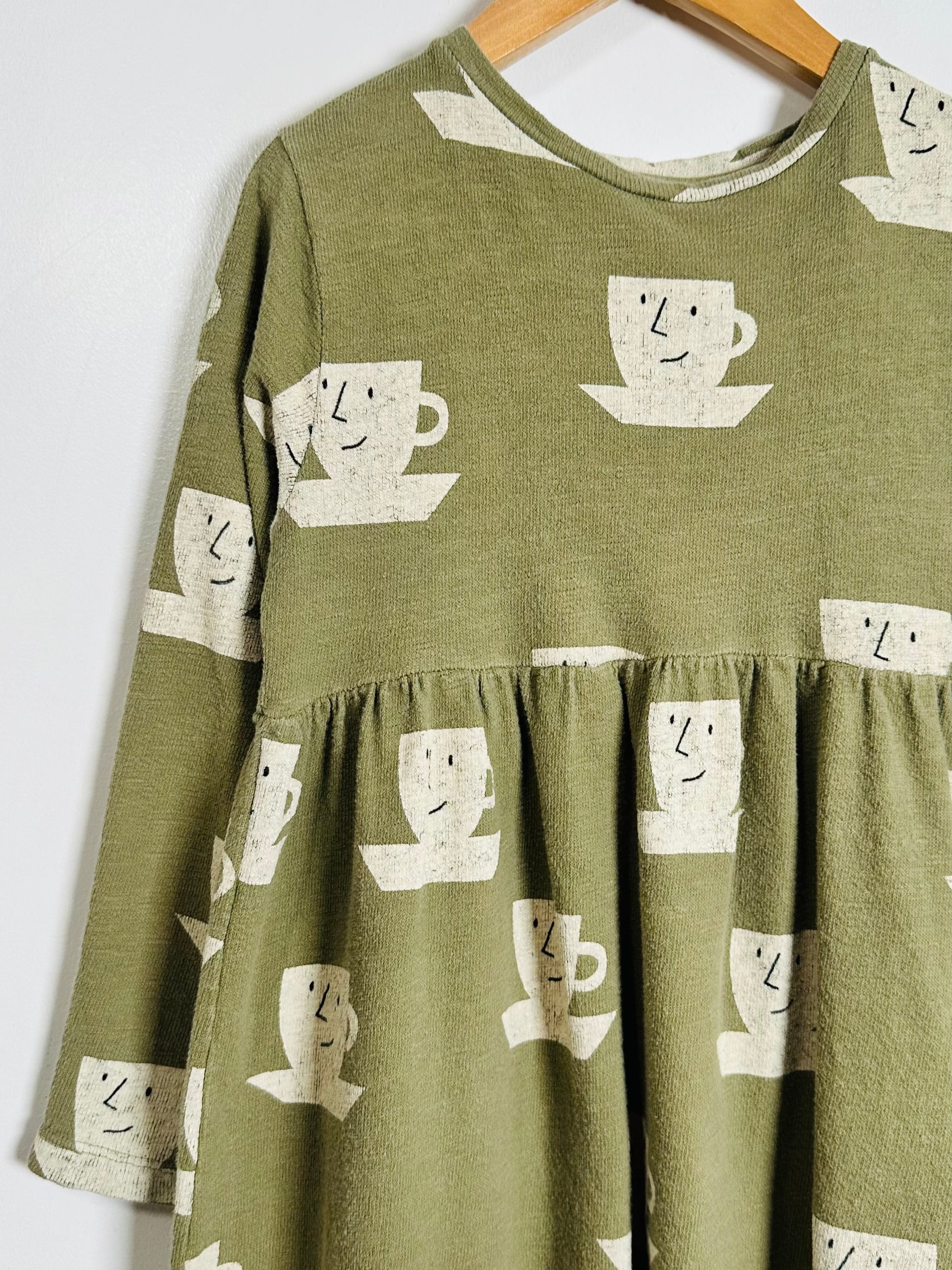 An olive green tiered long-sleeve dress with a white coffee cup pattern, hanging on a wooden hanger against a plain white wall.