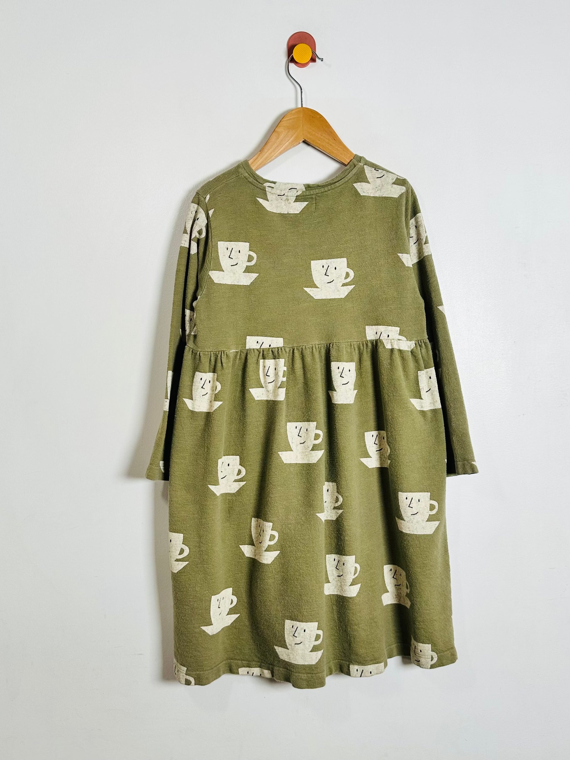 An olive green long-sleeve dress with a white coffee cup pattern, hanging on a wooden hanger against a plain white wall.