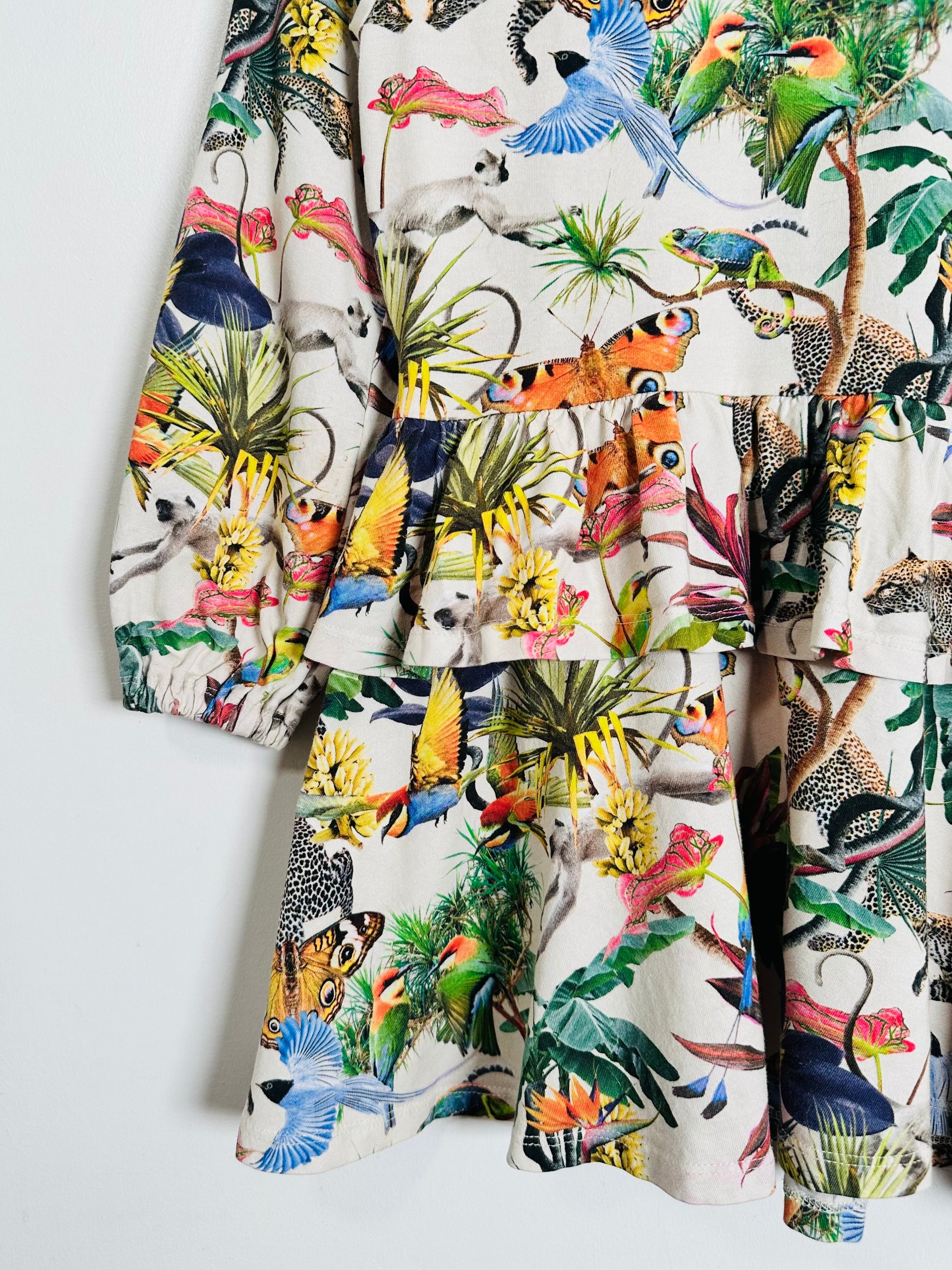 A tiered ruffle dress featuring a vibrant, multicolor jungle and animal print on a cream background, with long sleeves and a ruffled waist.
