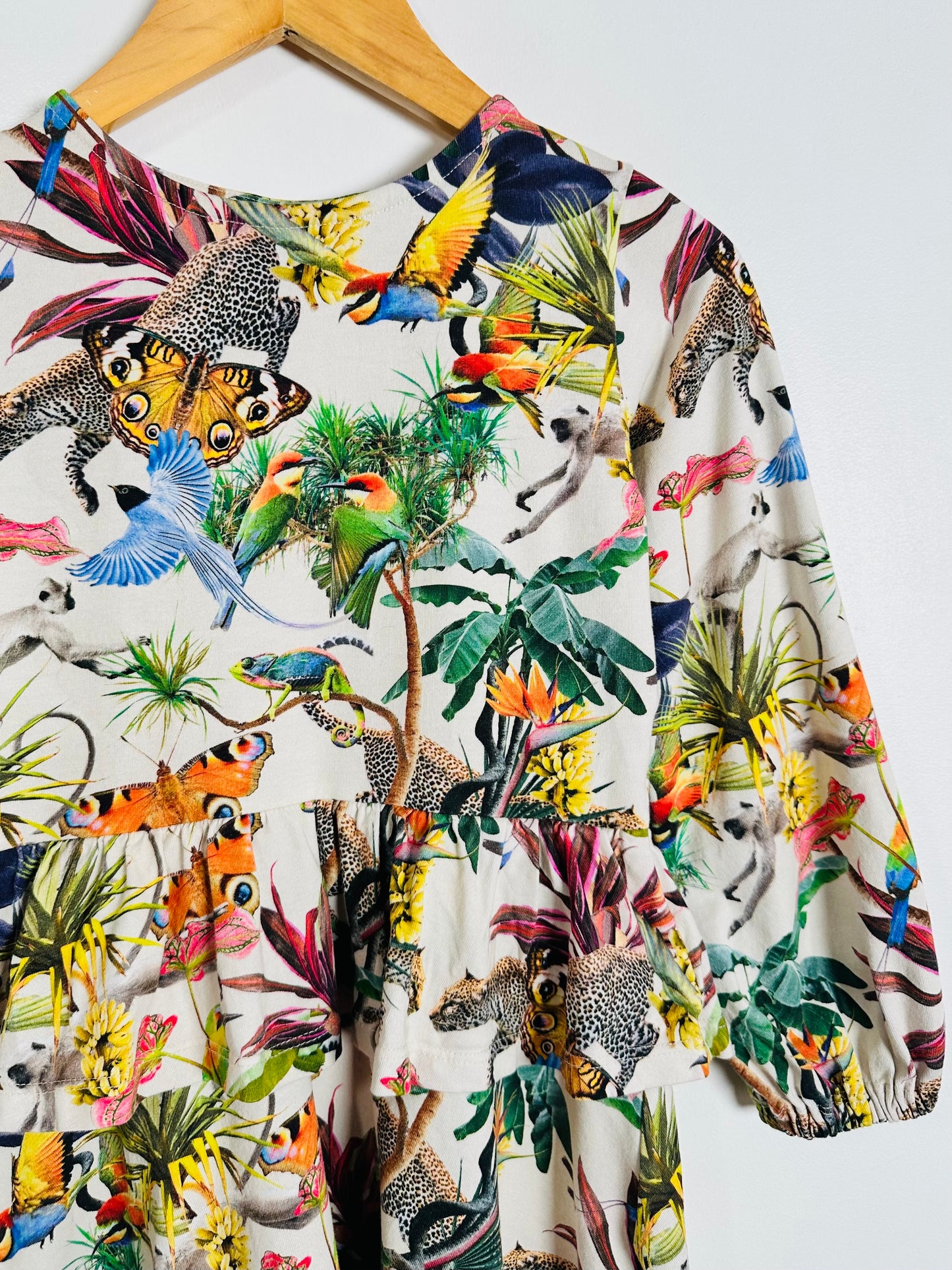A long-sleeve top with a vibrant multicolor jungle print featuring toucans, parrots, monkeys, and tropical plants on an off-white background, hanging on a wooden hanger against a plain white wall.