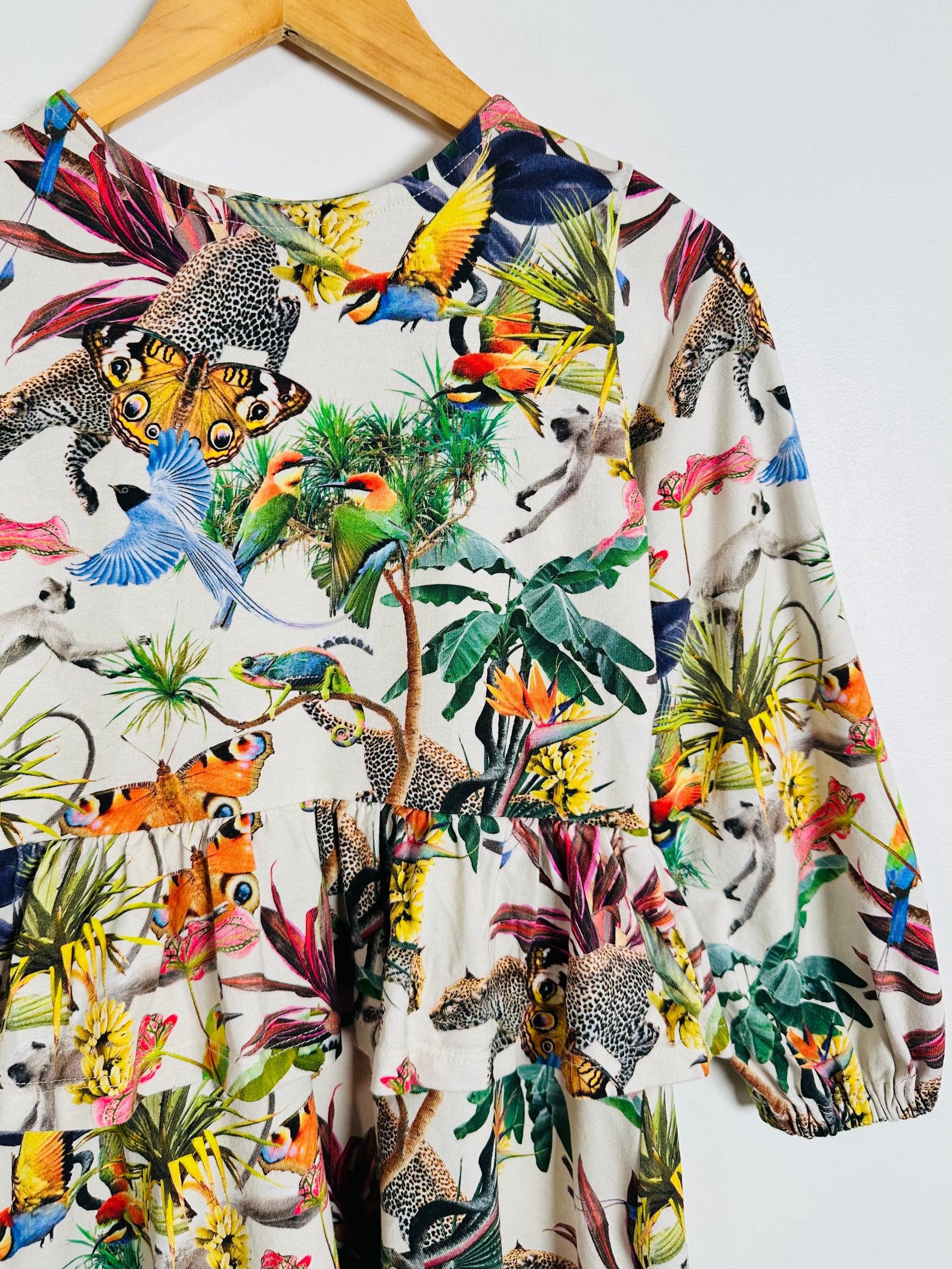 A long-sleeve top with a vibrant multicolor jungle print featuring toucans, parrots, monkeys, and tropical plants on an off-white background, hanging on a wooden hanger against a plain white wall.