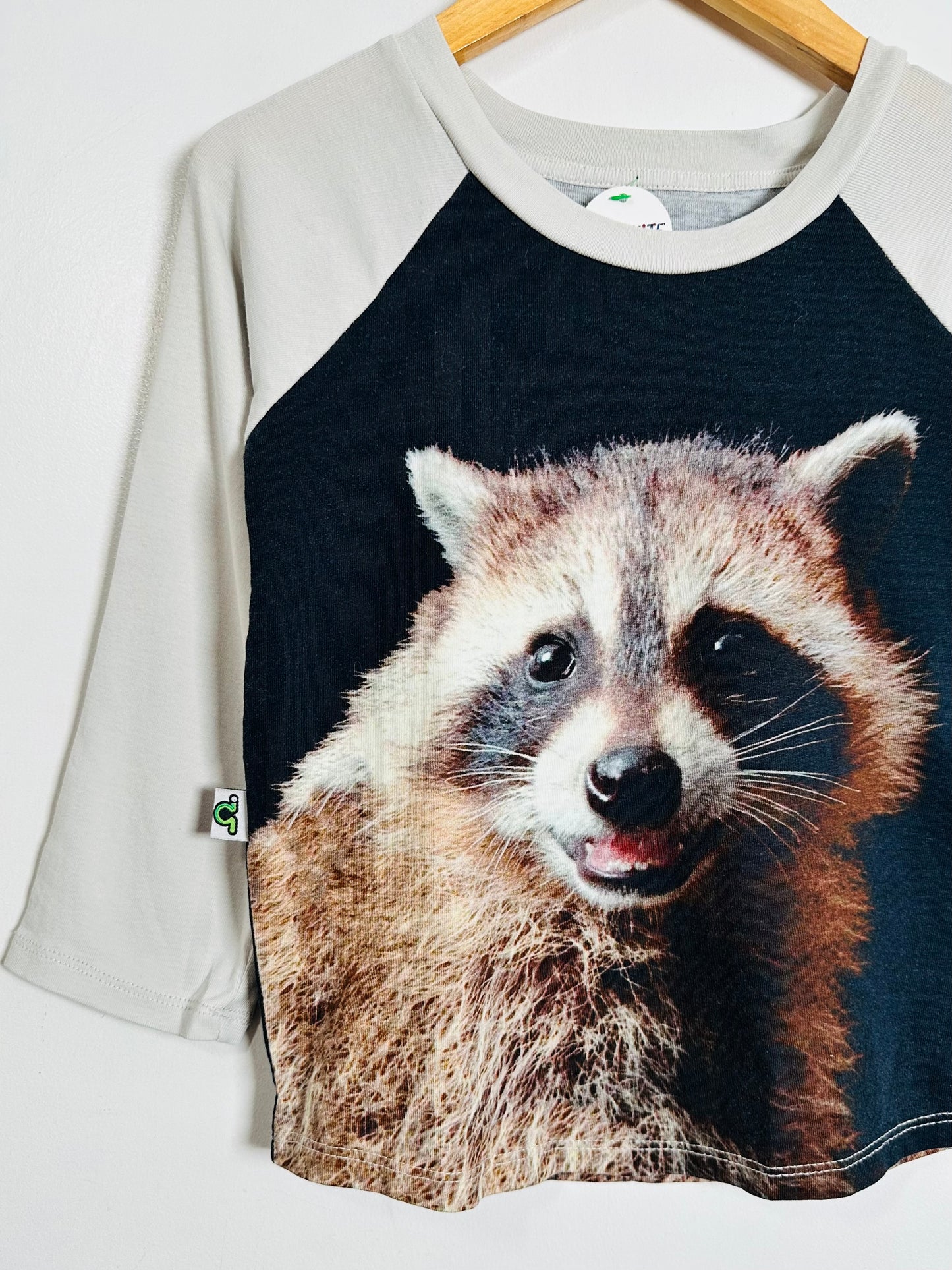 A raglan-style t-shirt with a large printed raccoon face on the front, featuring a black body and light gray sleeves, hanging on a wooden hanger against a white wall.
