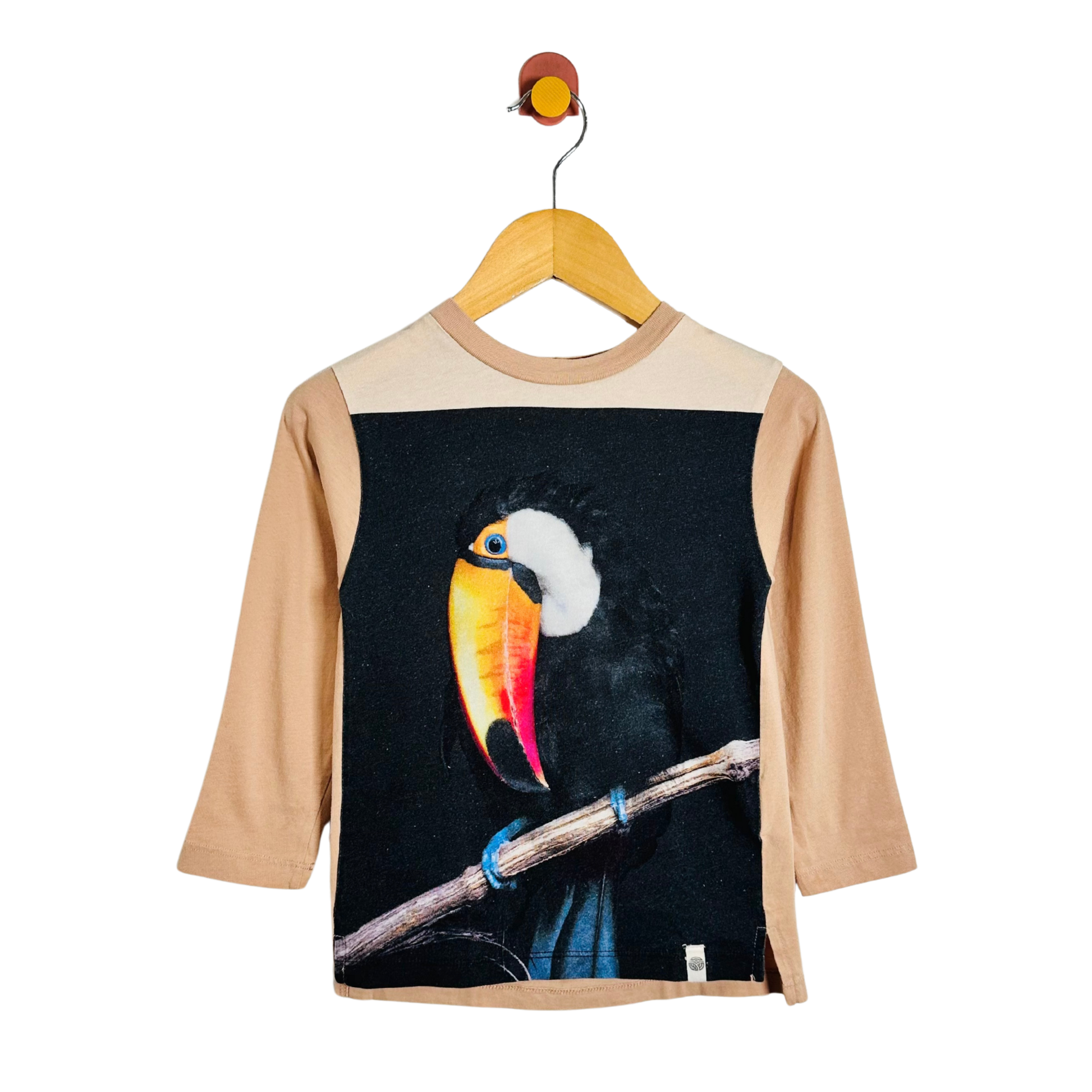 A children's long-sleeve t-shirt with beige sleeves and shoulder yoke, featuring a large graphic print of a toucan on the front over a black background. The shirt is displayed on a wooden hanger against a plain white background.