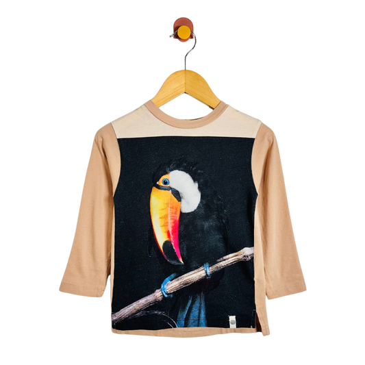A children's long-sleeve t-shirt with beige sleeves and shoulder yoke, featuring a large graphic print of a toucan on the front over a black background. The shirt is displayed on a wooden hanger against a plain white background.
