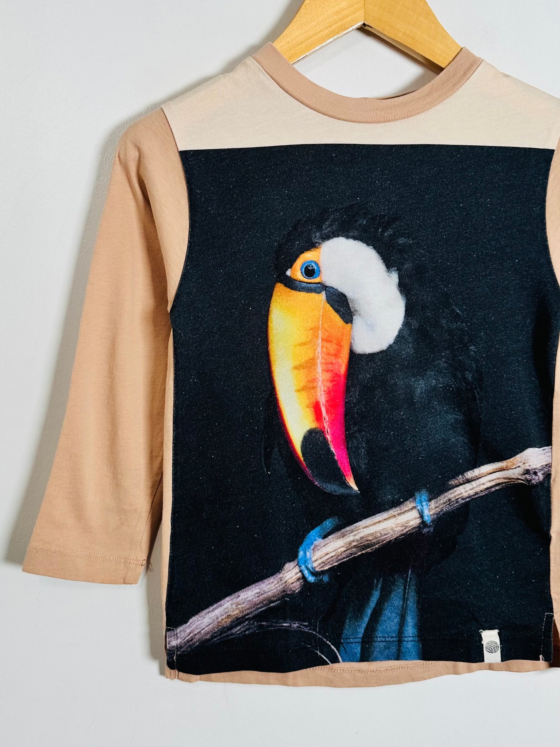 A children's long-sleeve shirt with beige sleeves and collar, featuring a large front graphic print of a toucan with a bright yellow, orange, and red beak on a black background, hanging on a wooden hanger against a light gray wall.