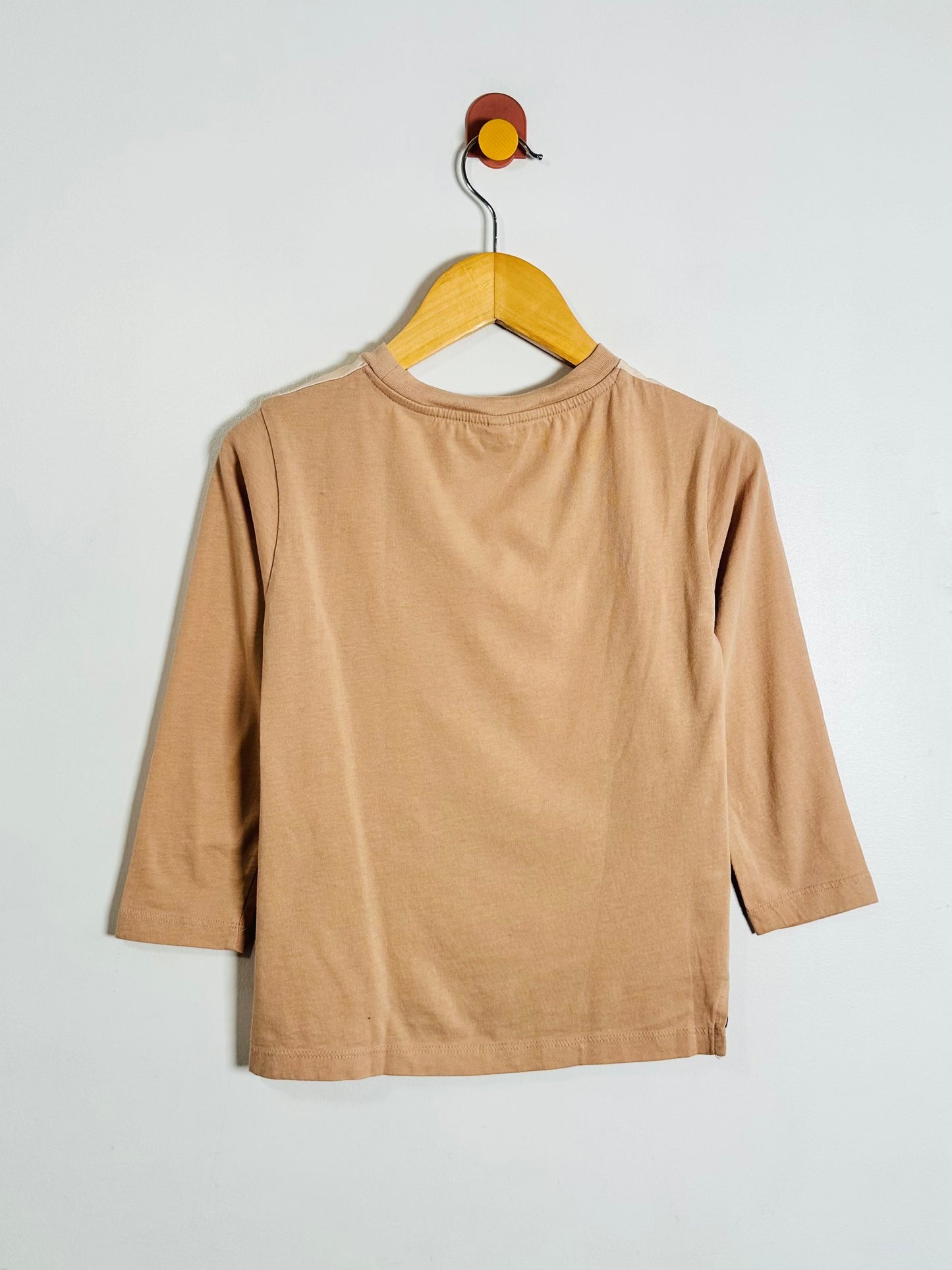 A light brown long-sleeve t-shirt hanging on a wooden hanger against a plain light gray wall.