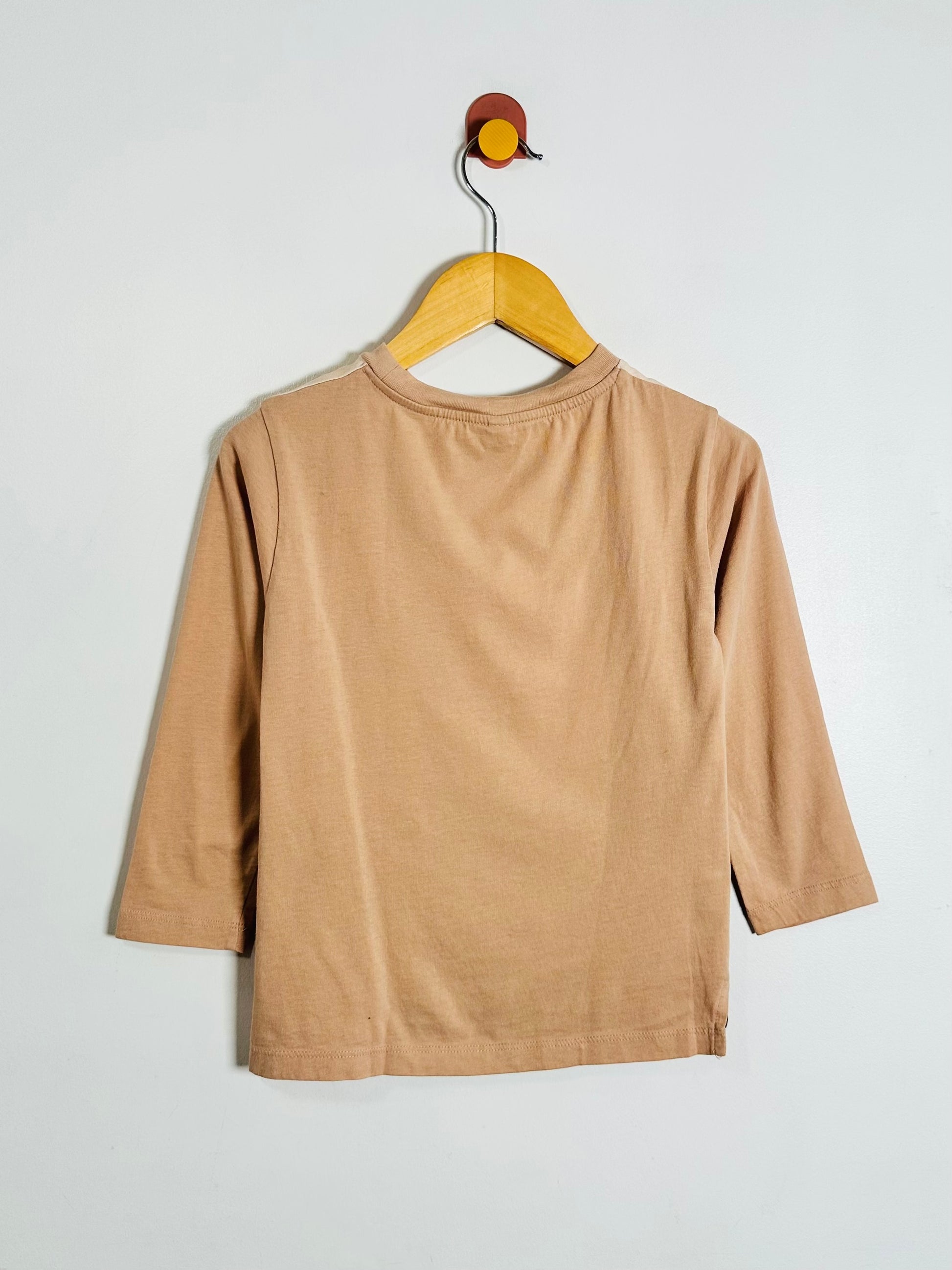 A light brown long-sleeve t-shirt hanging on a wooden hanger against a plain light gray wall.