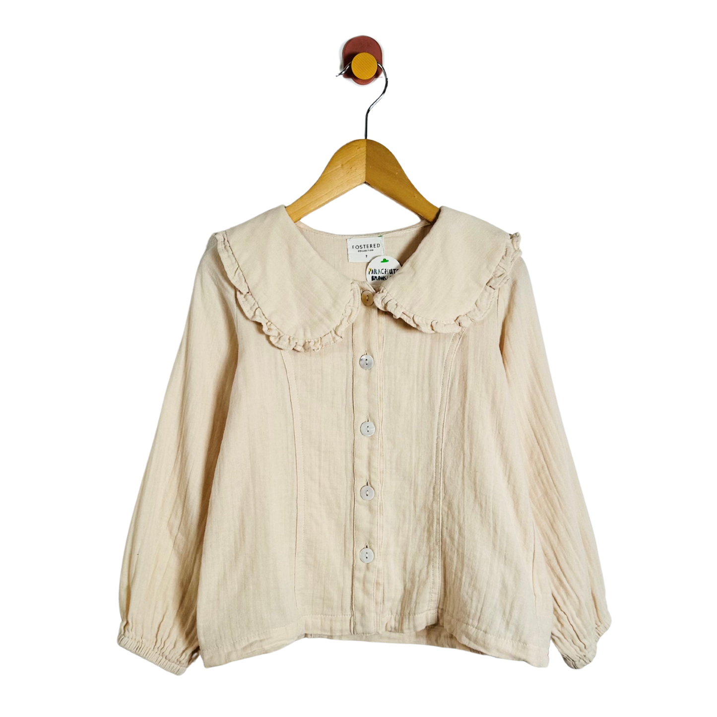 A cream-colored crinkle gauze blouse with a ruffled Peter Pan collar and button-front closure, hanging on a wooden hanger against a plain white background.