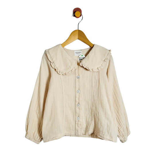 A cream-colored crinkle gauze blouse with a ruffled Peter Pan collar and button-front closure, hanging on a wooden hanger against a plain white background.