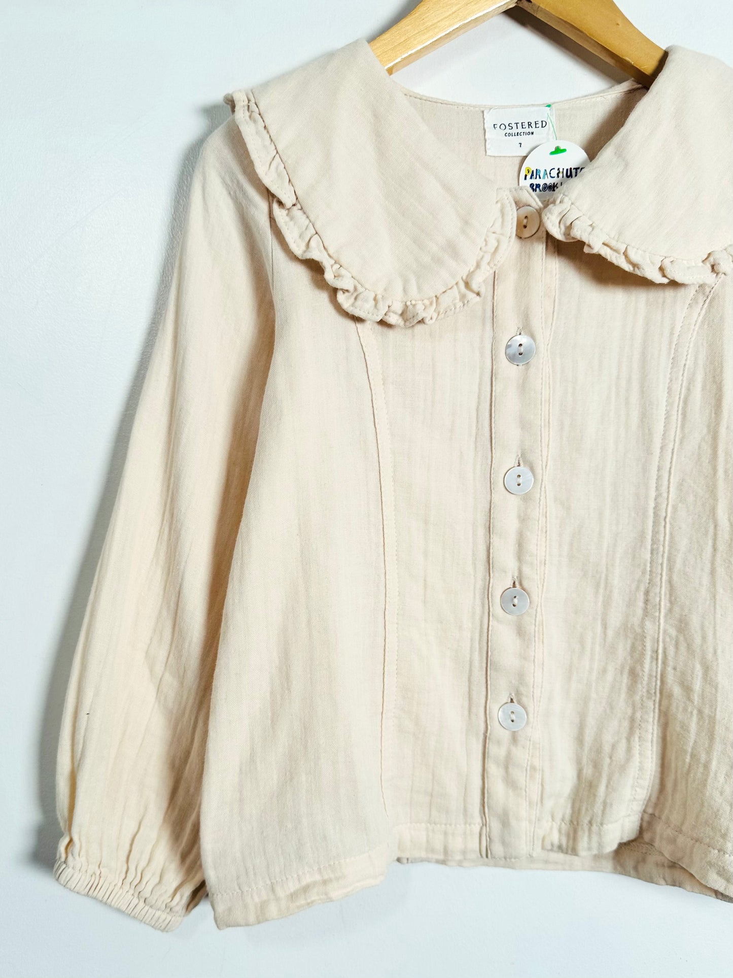 A cream-colored cotton blouse with a ruffled Peter Pan collar and button-front closure, hanging on a wooden hanger against a plain white background.
