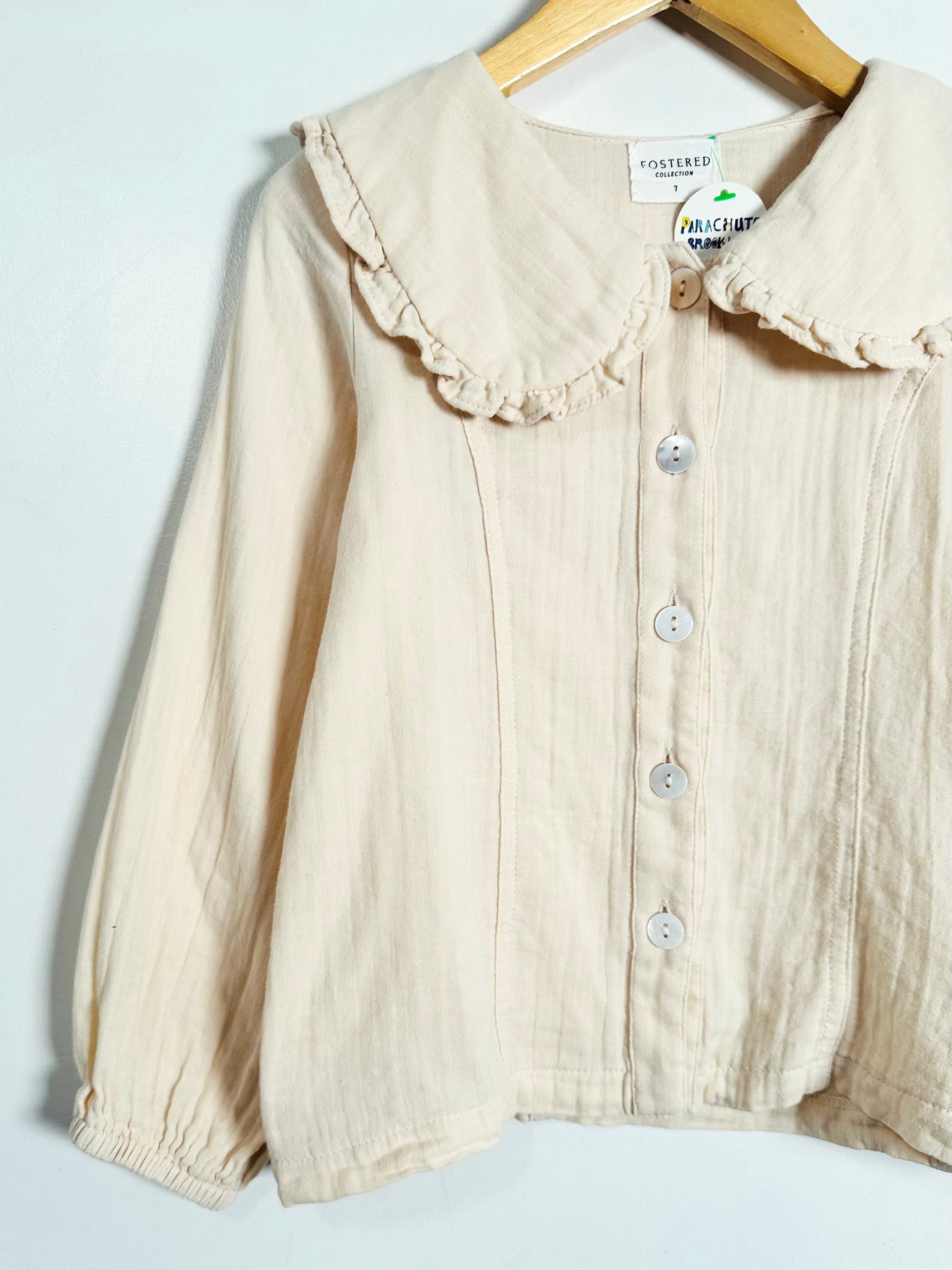 A cream-colored cotton blouse with a ruffled Peter Pan collar and button-front closure, hanging on a wooden hanger against a plain white background.