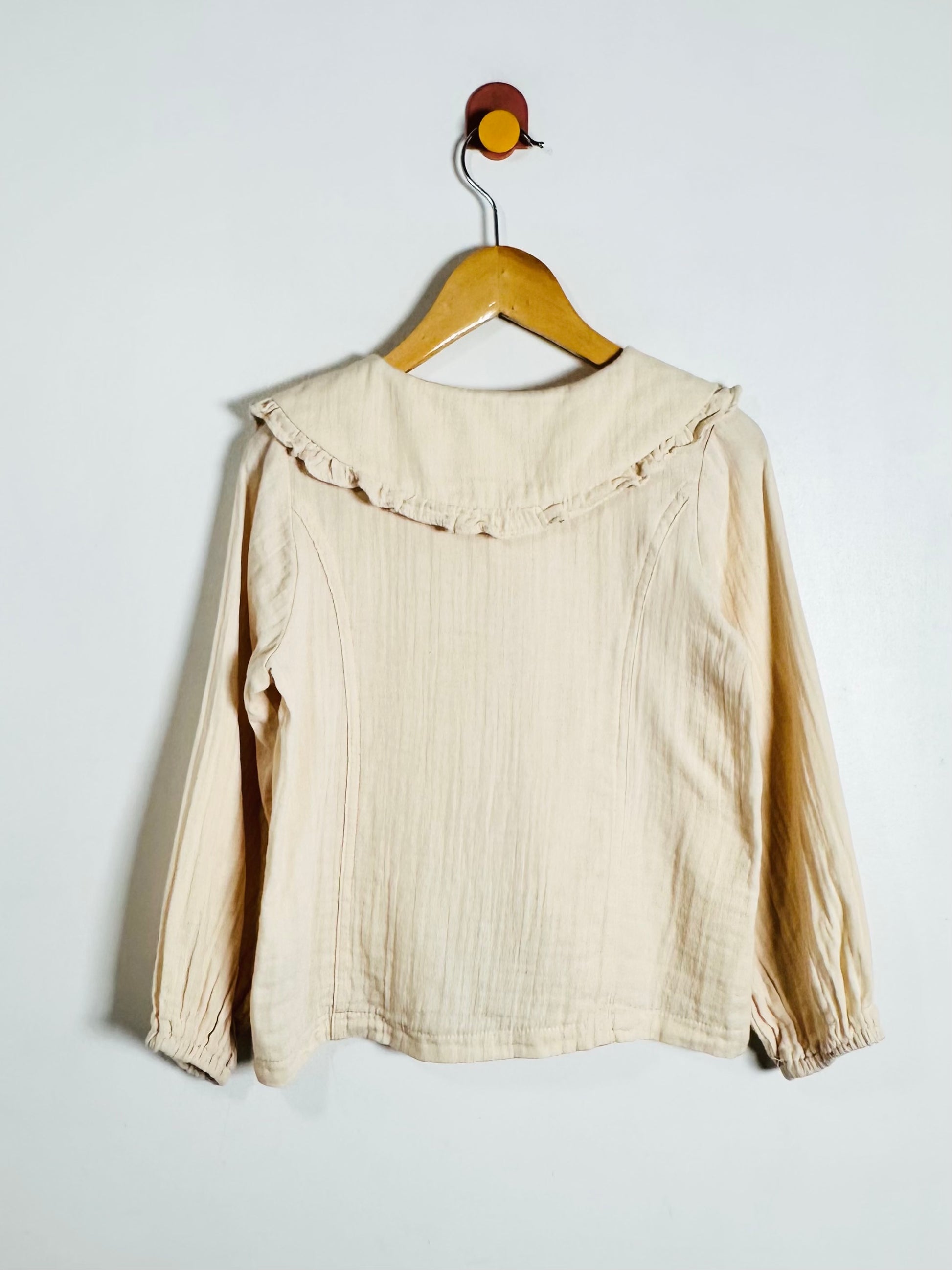 A beige long-sleeve blouse with a ruffled collar, hanging on a wooden hanger against a plain white wall.