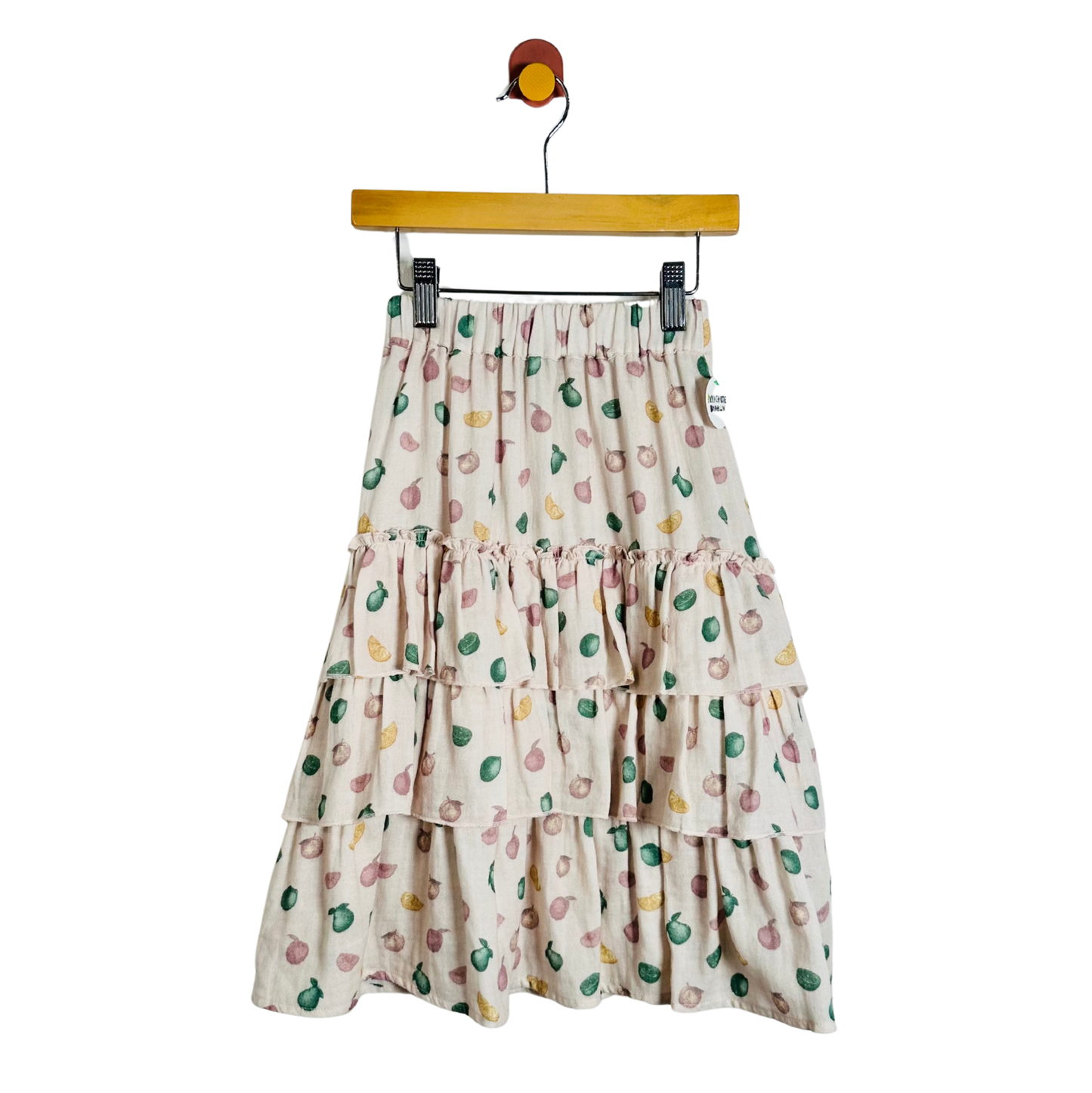 A tiered midi skirt with a colorful fruit print on a cream background, hanging from a wooden hanger against a plain white background.
