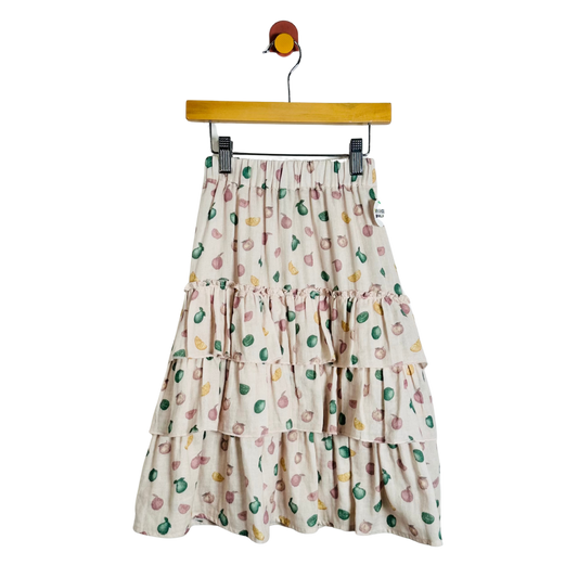 A tiered midi skirt with a colorful fruit print on a cream background, hanging from a wooden hanger against a plain white background.