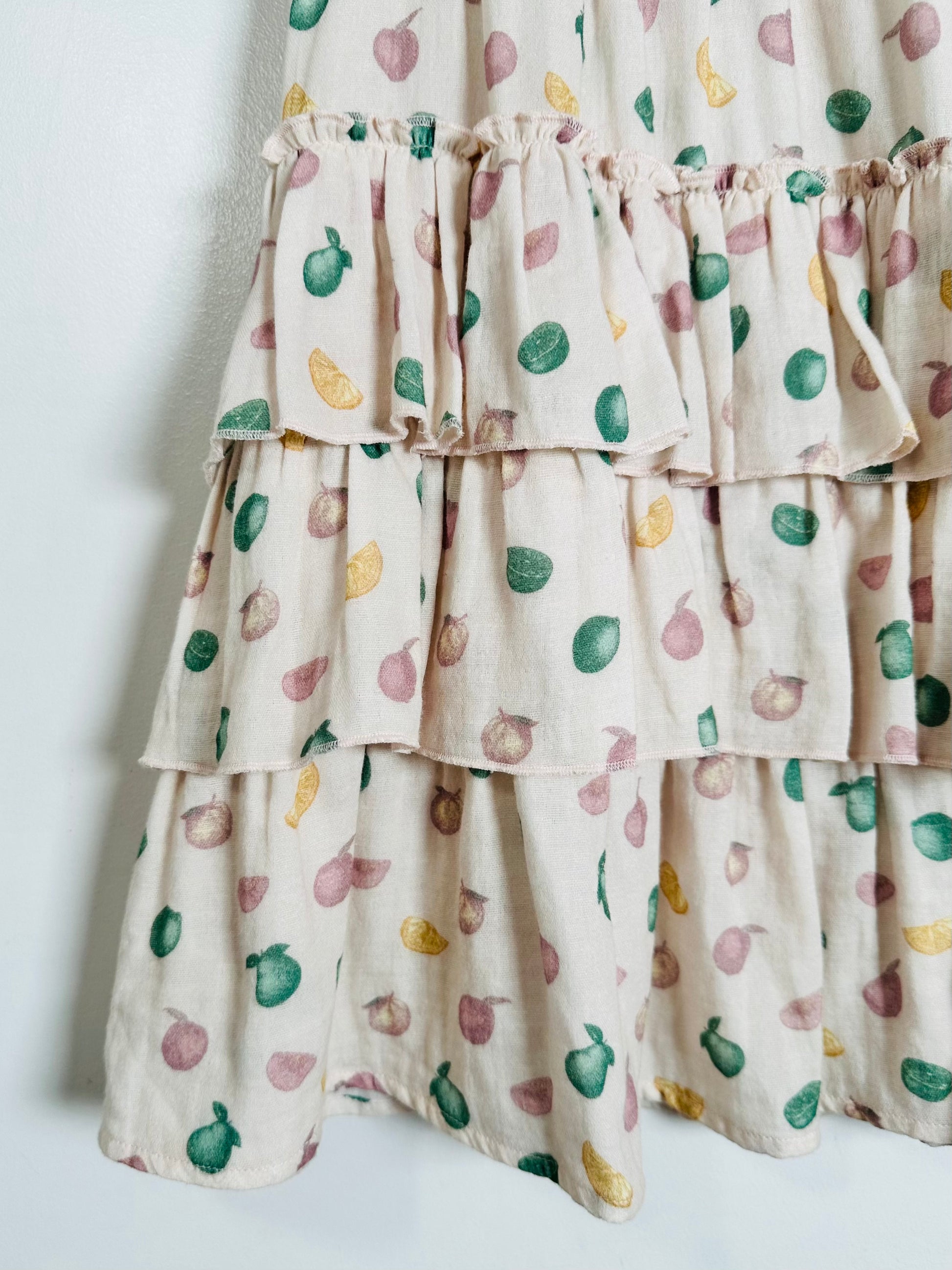 Close-up of a tiered ruffle dress or skirt in a warm cream fabric with a scattered print of green pears, pinkish-purple onions, and yellow citrus slices.