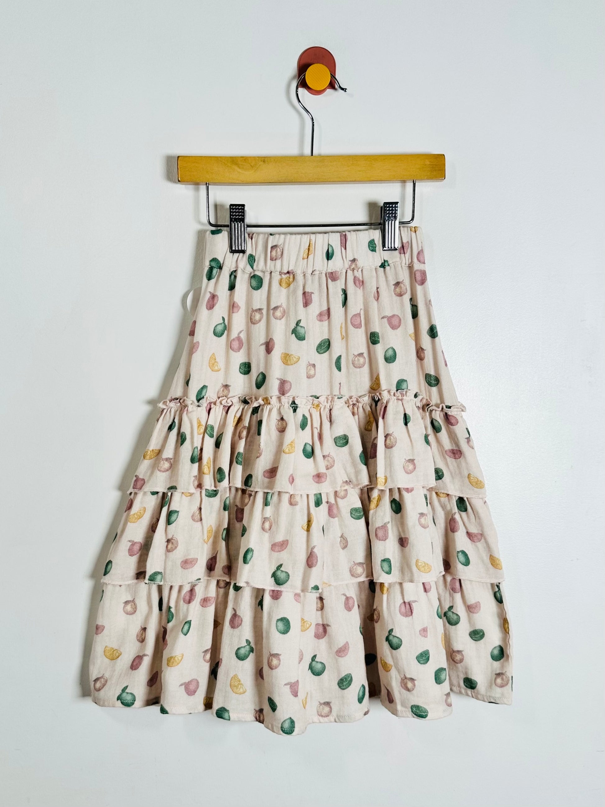 A tiered midi skirt with a beige base and a colorful pattern of small green, pink, and yellow abstract shapes, hanging from a wooden hanger against a plain white wall.