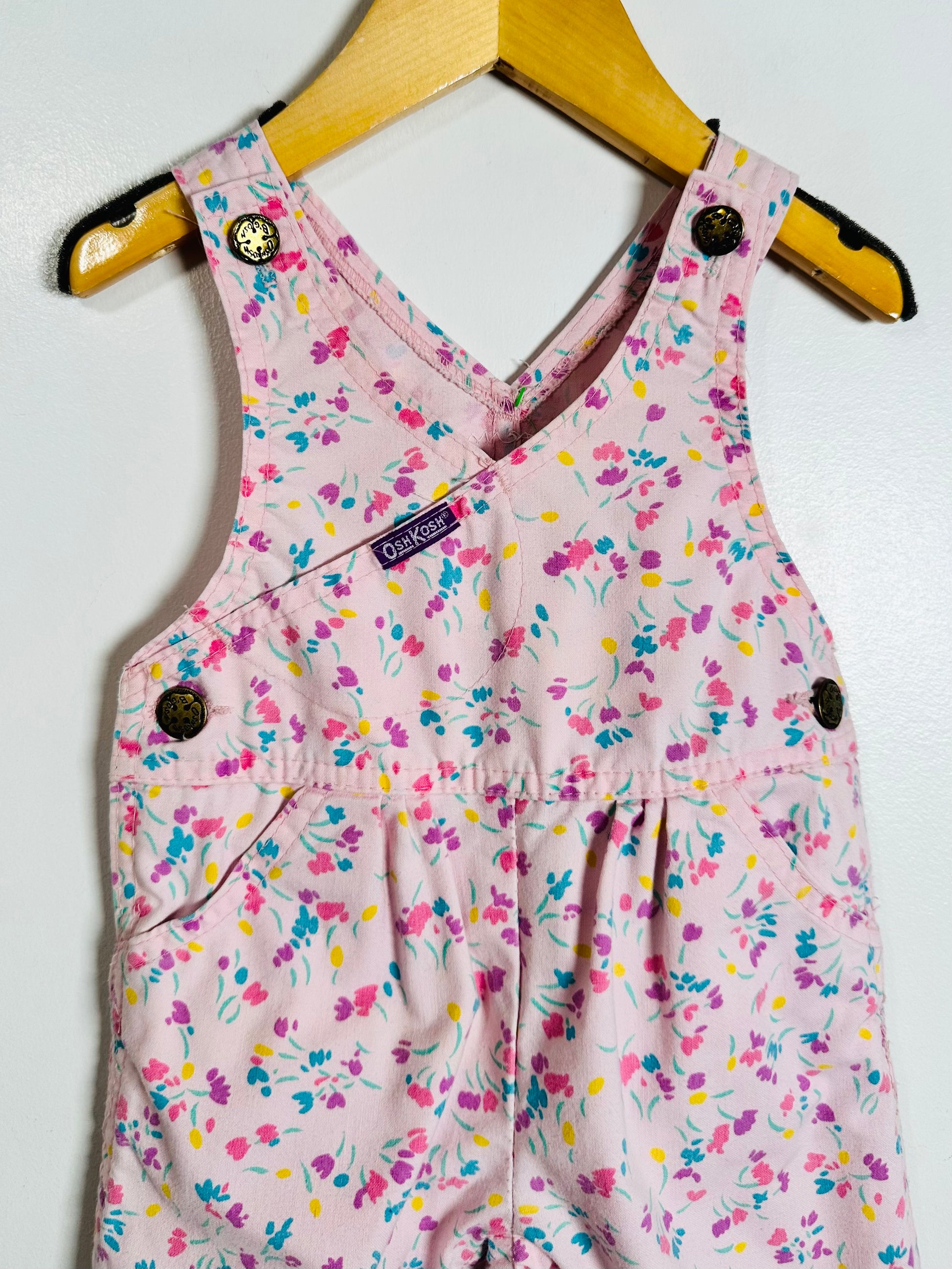 A baby girl's floral print overall romper in light pink, hanging on a wooden hanger against a white wall. The romper features a delicate pink, purple, blue, and yellow floral pattern, a wrap-style front, and dark brown snap buttons.