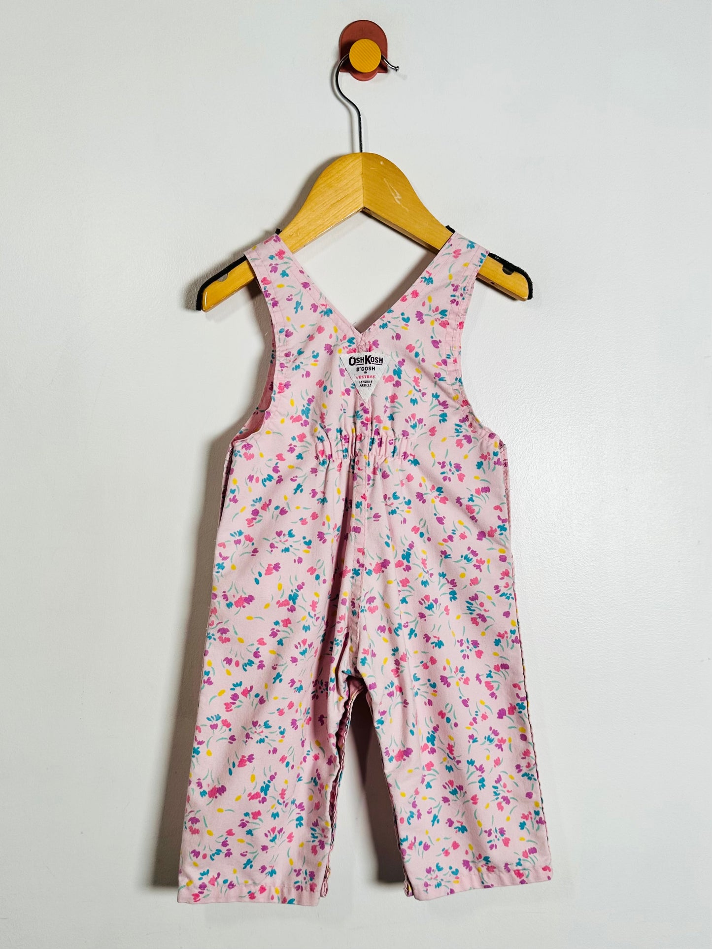 A baby girl's sleeveless floral jumpsuit in pink, featuring a delicate multicolor floral pattern, hanging on a wooden hanger against a plain white wall.