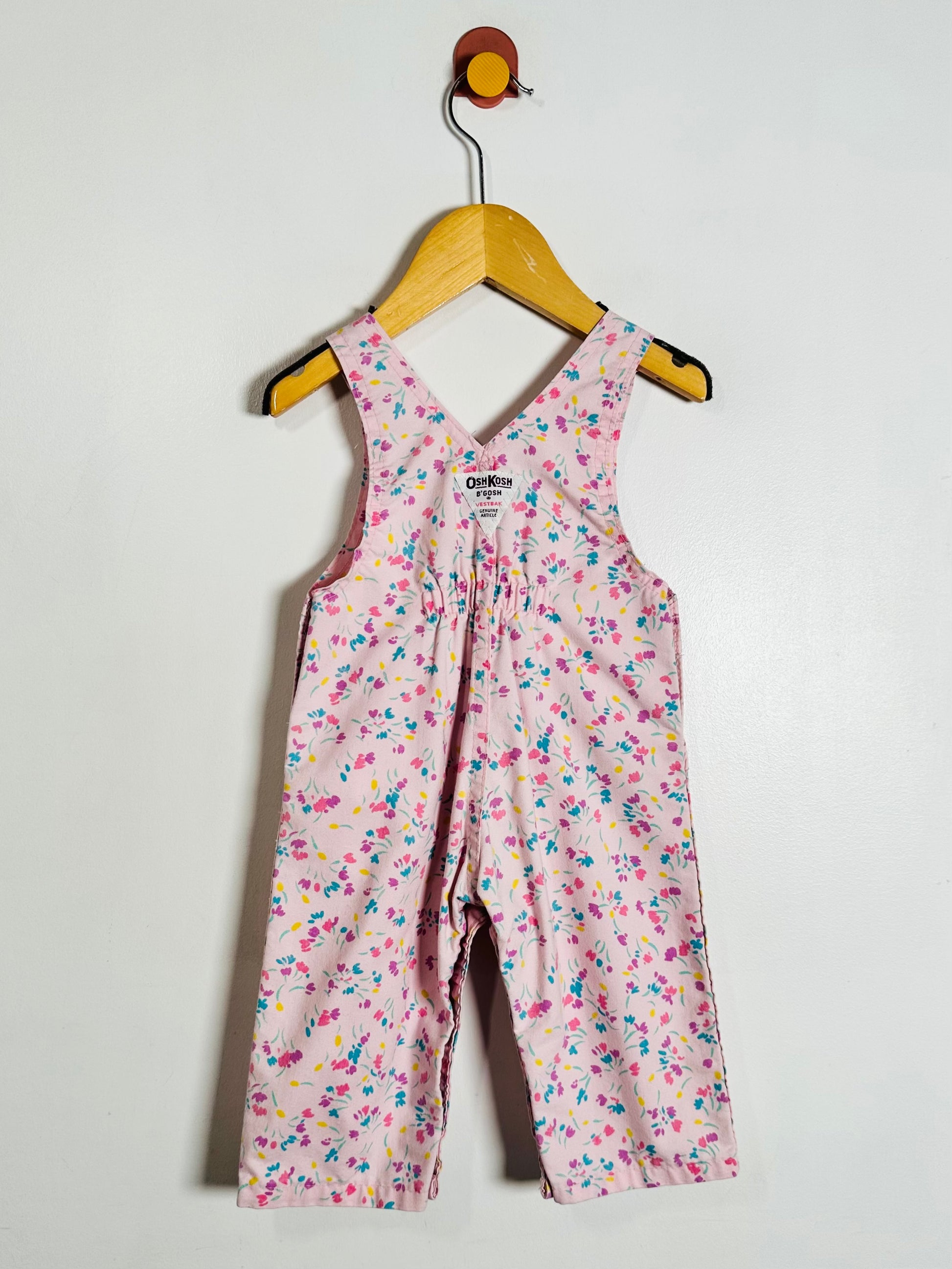 A baby girl's sleeveless floral jumpsuit in pink, featuring a delicate multicolor floral pattern, hanging on a wooden hanger against a plain white wall.