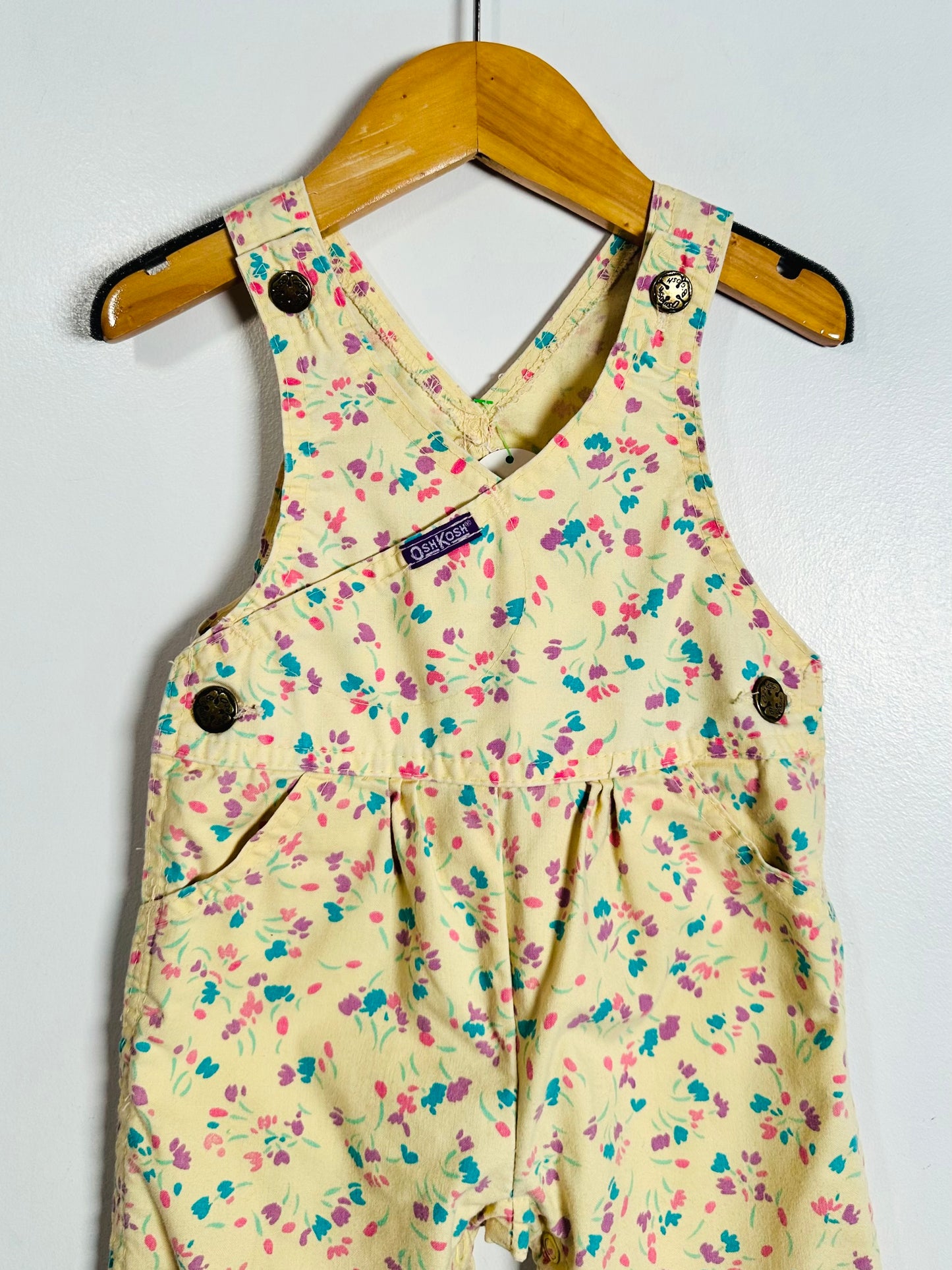 A baby romper in a pale yellow floral print, hanging on a wooden hanger against a white wall. The floral pattern features pink, purple, and blue flowers with green stems.
