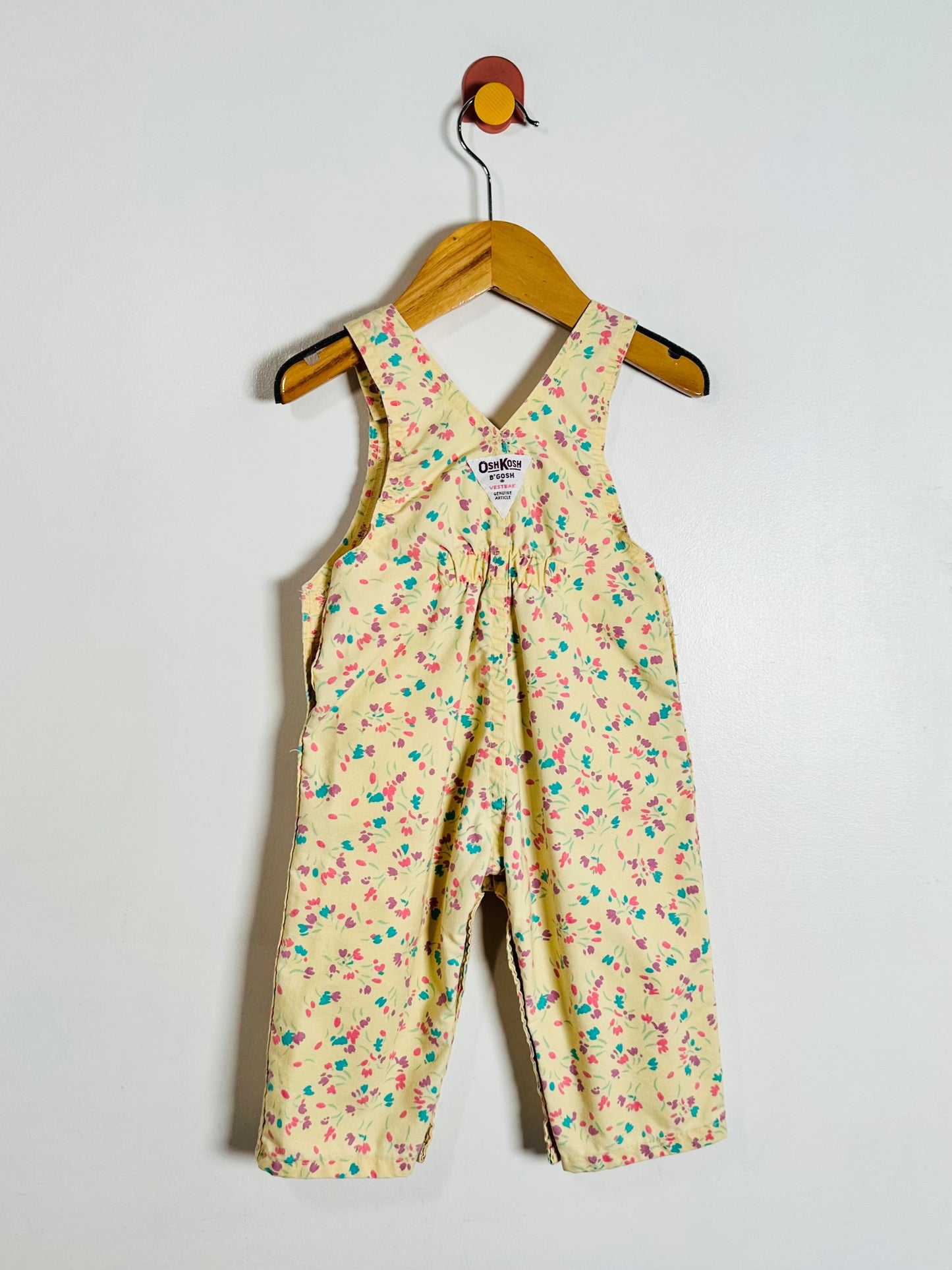 A baby romper in a light yellow floral print, hanging on a wooden hanger against a plain white wall.