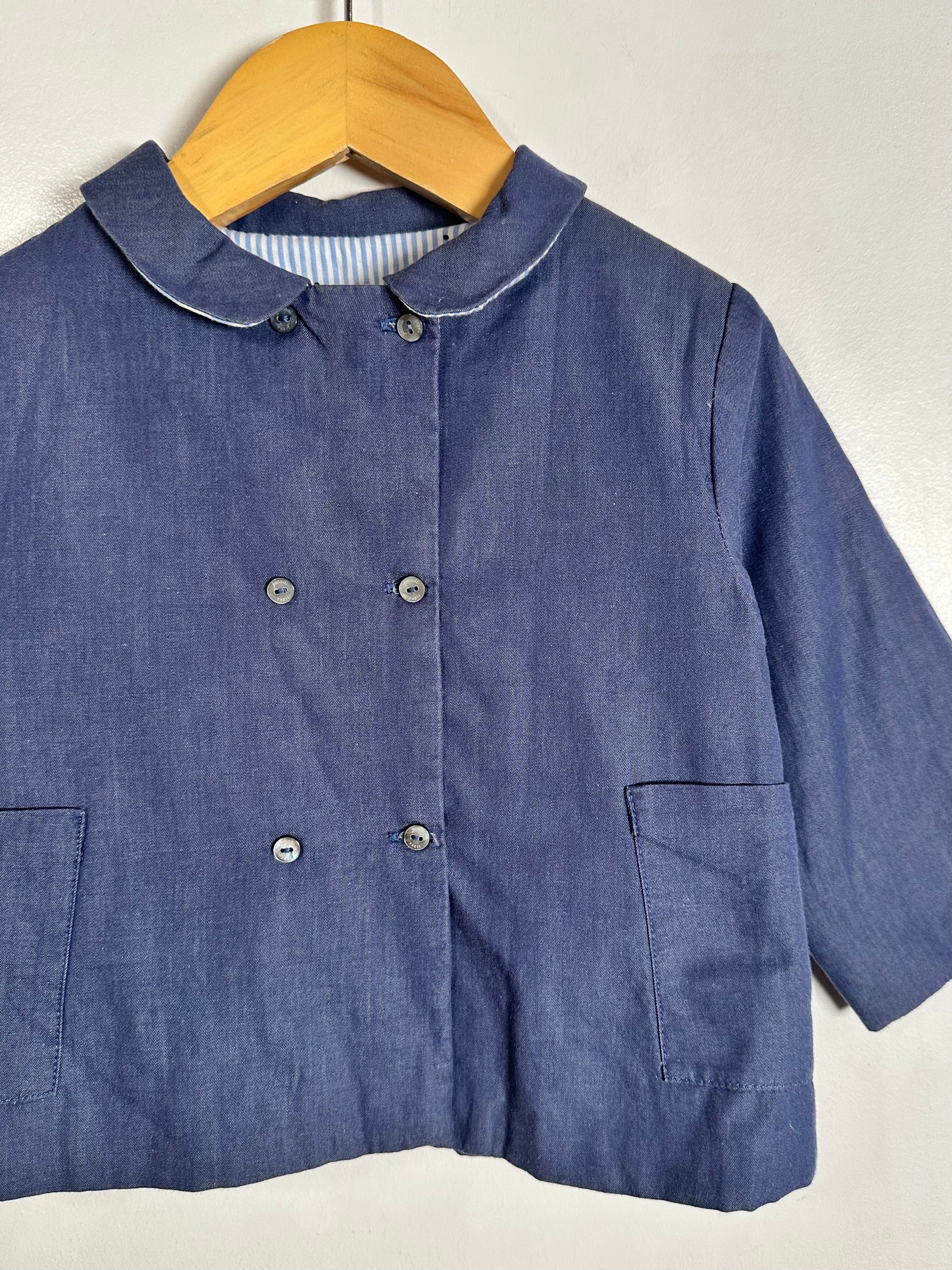 A blue denim-like children's jacket with a rounded collar, double-breasted buttons, and front patch pockets, hanging on a wooden hanger against a plain light wall.
