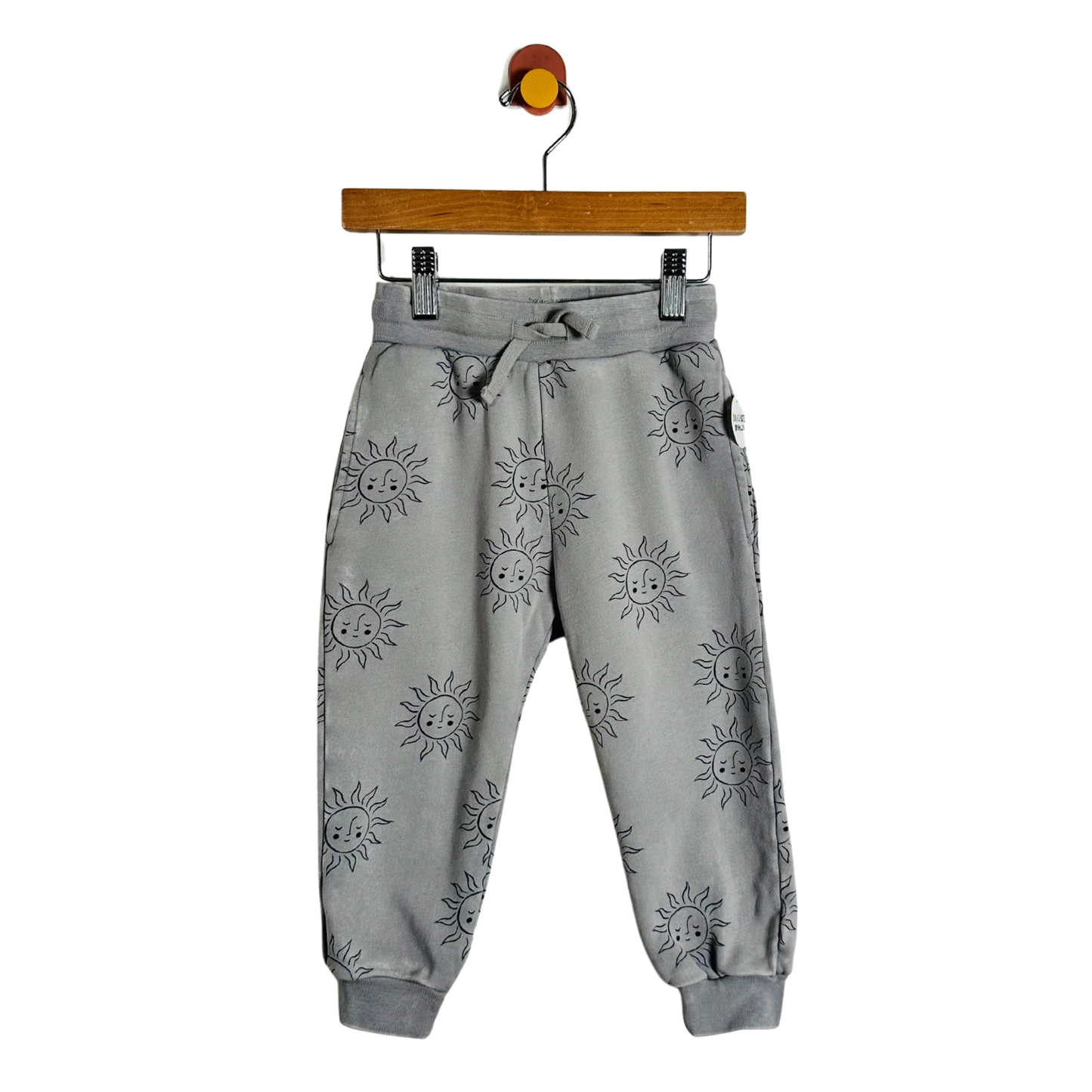 Gray jogger pants with a black sun print, hanging from a wooden hanger with a colorful hook against a plain white background.