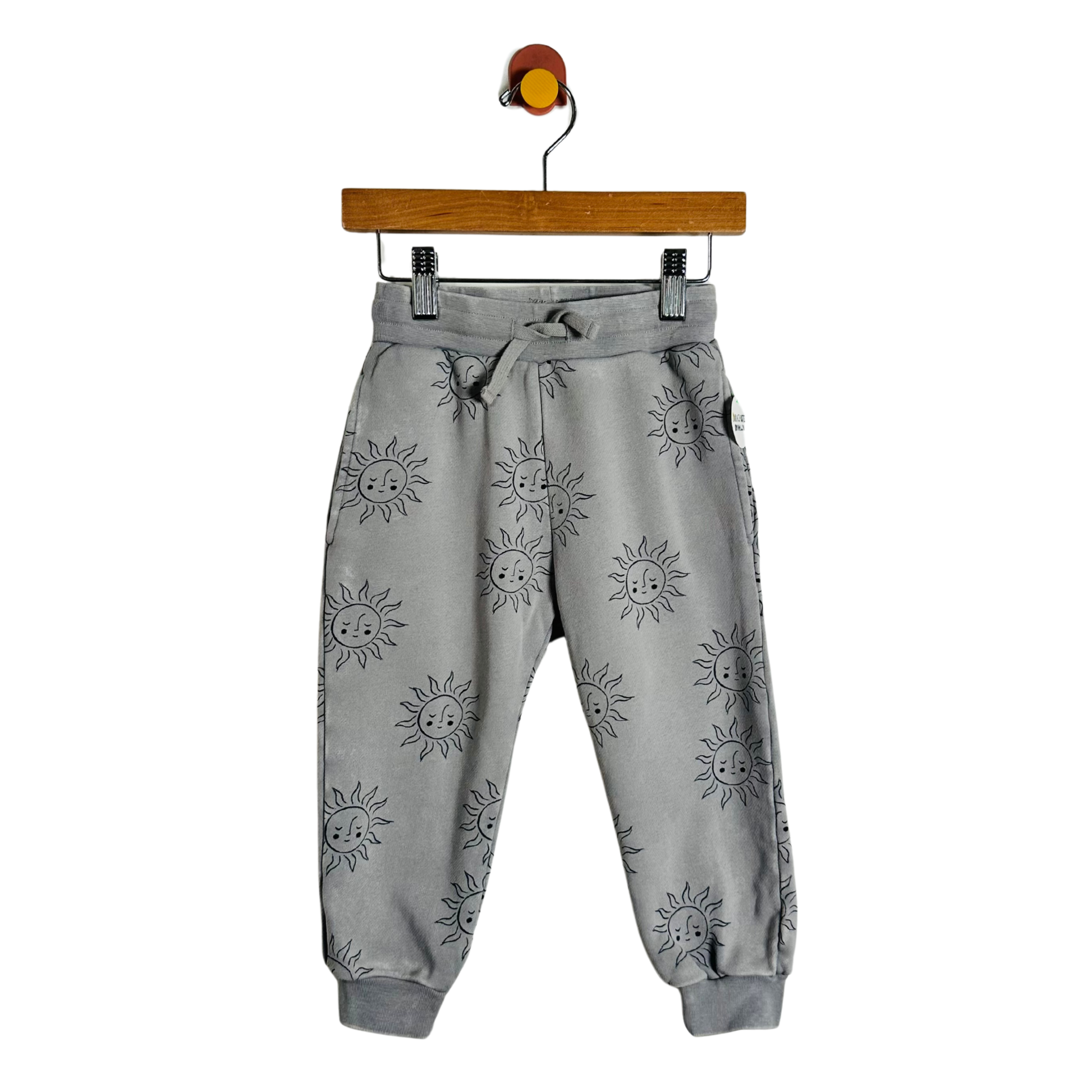 Gray jogger pants with a black sun print, hanging from a wooden hanger with a colorful hook against a plain white background.