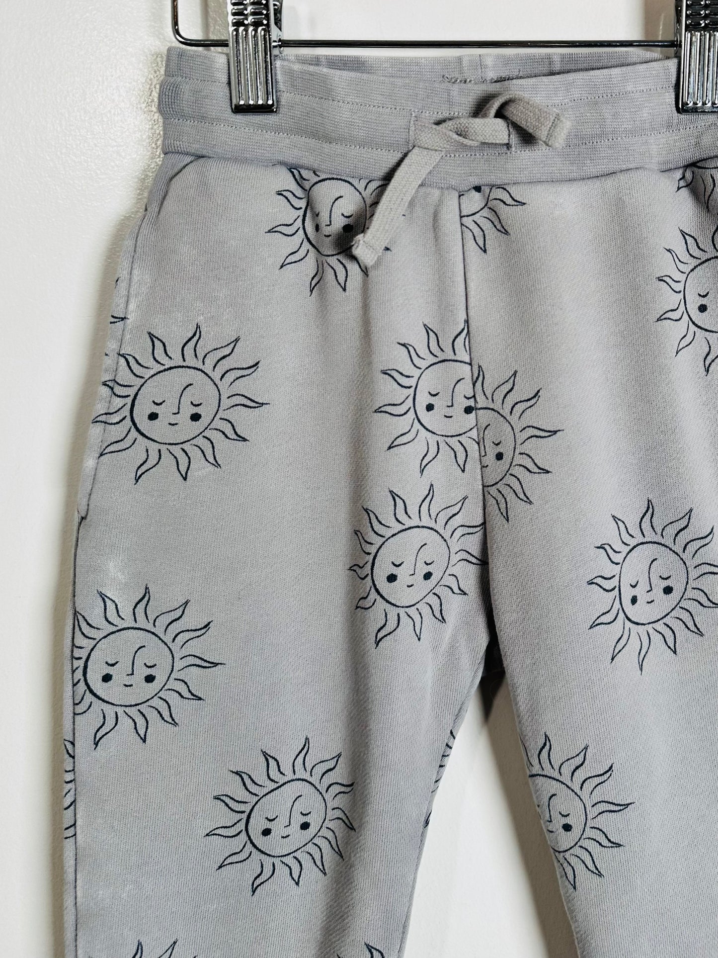 Light gray sweatpants with an all-over black line-art print of sun faces, hanging from a black metal hanger against a plain white wall.