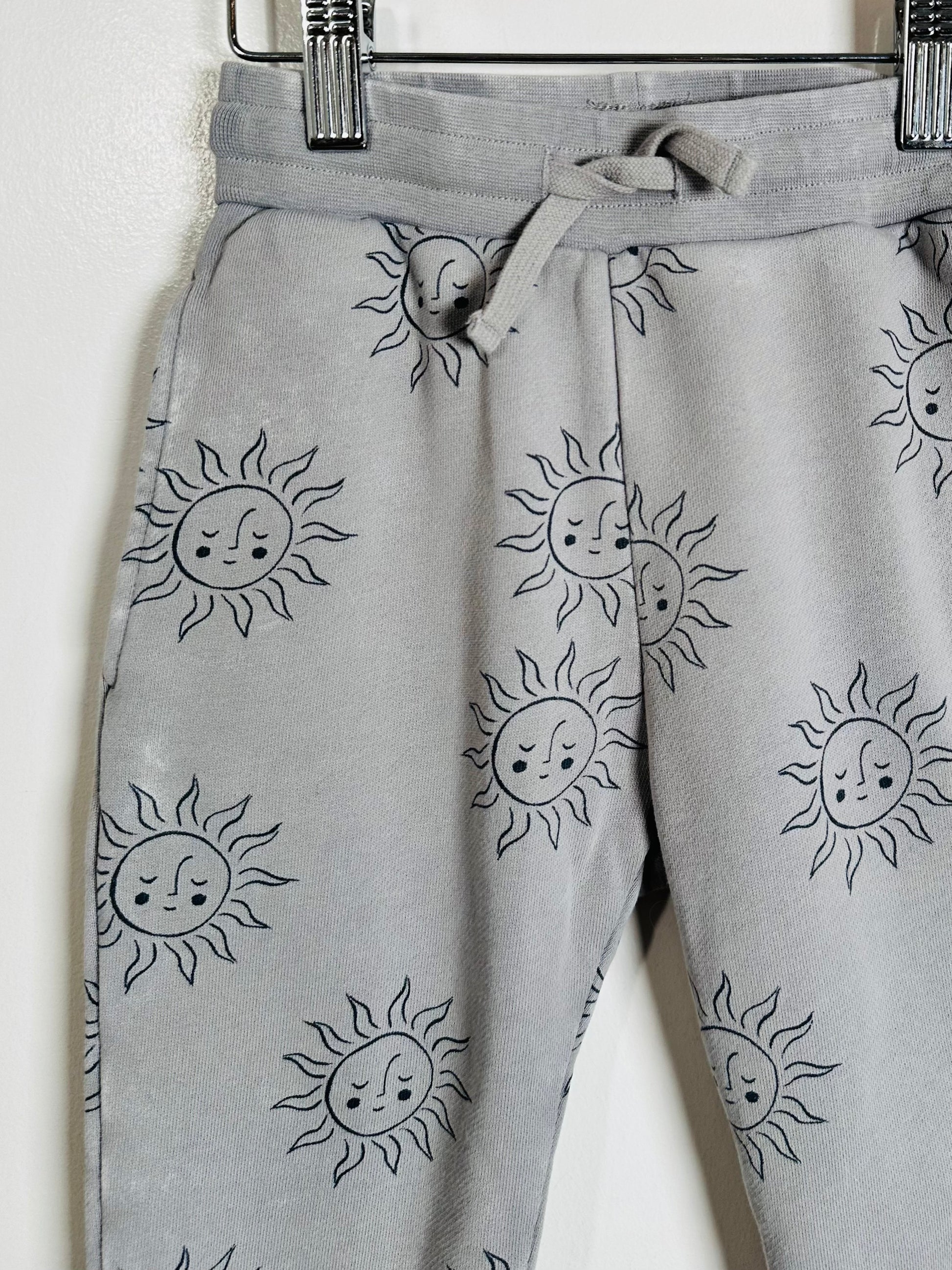 Light gray sweatpants with an all-over black line-art print of sun faces, hanging from a black metal hanger against a plain white wall.