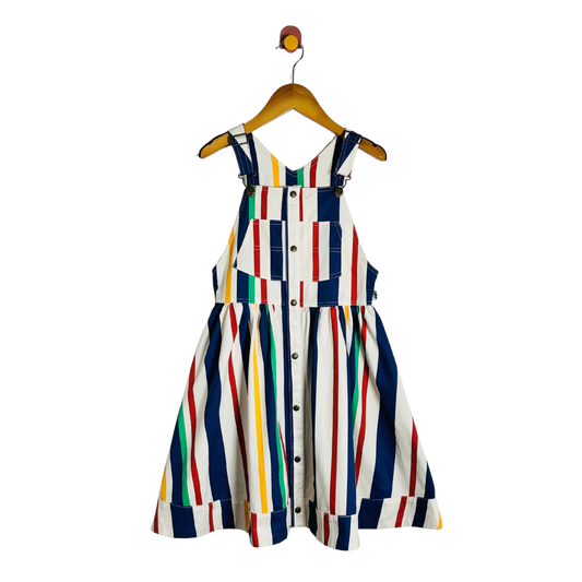 A sleeveless children's dress with a white base and vertical multicolor stripes in navy, red, yellow, green, and orange, hanging on a wooden hanger against a plain white background.