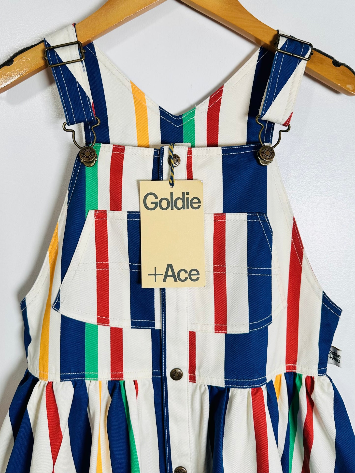 A children's striped overall dress hanging on a wooden hanger against a white wall. The dress has vertical multicolor stripes in blue, red, green, yellow, and white, with a bib-style neckline, front pockets, and a pleated skirt.