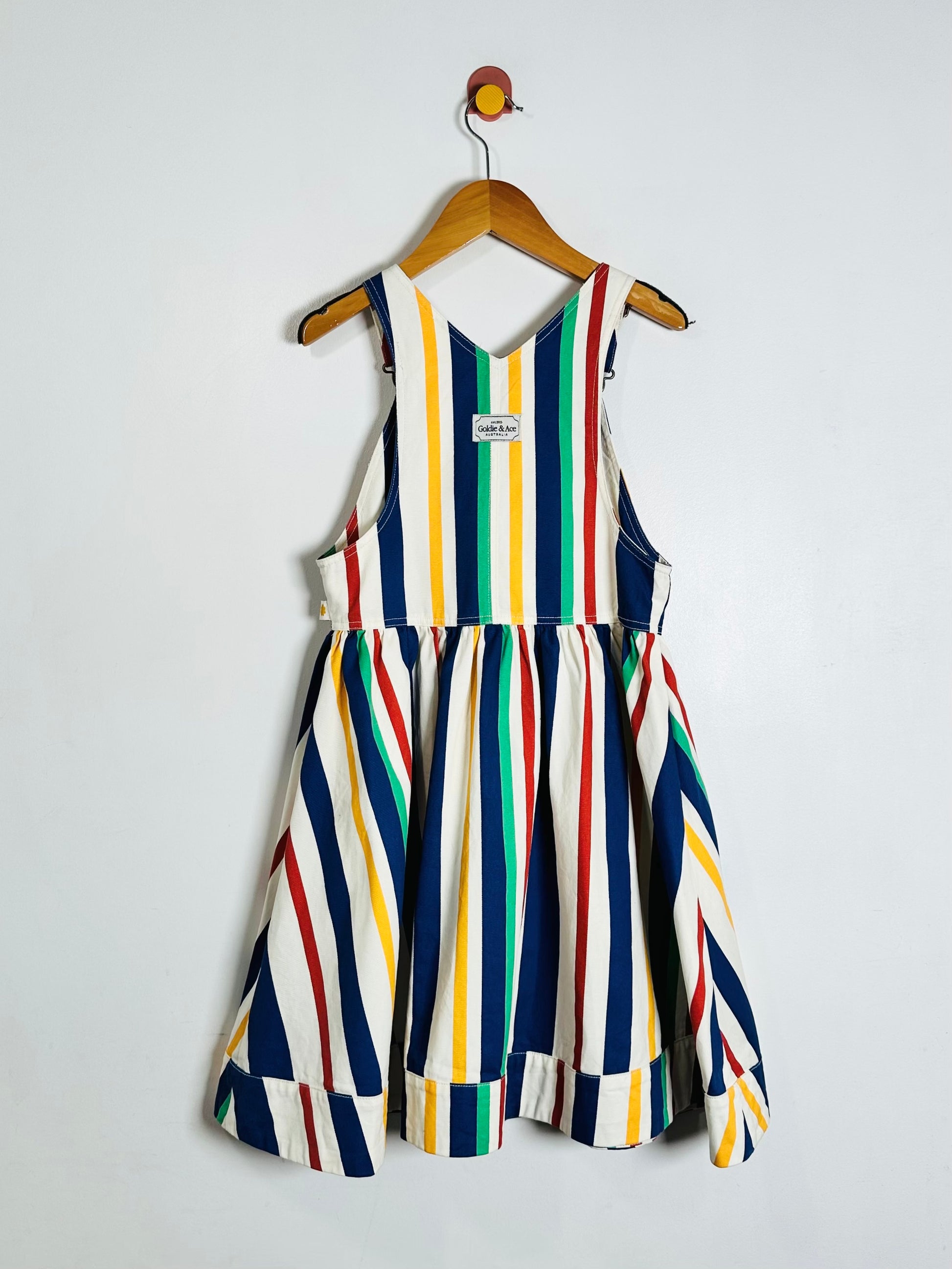 A sleeveless dress with vertical multicolor stripes in navy, white, yellow, green, and red, hanging on a wooden hanger against a plain light gray wall.