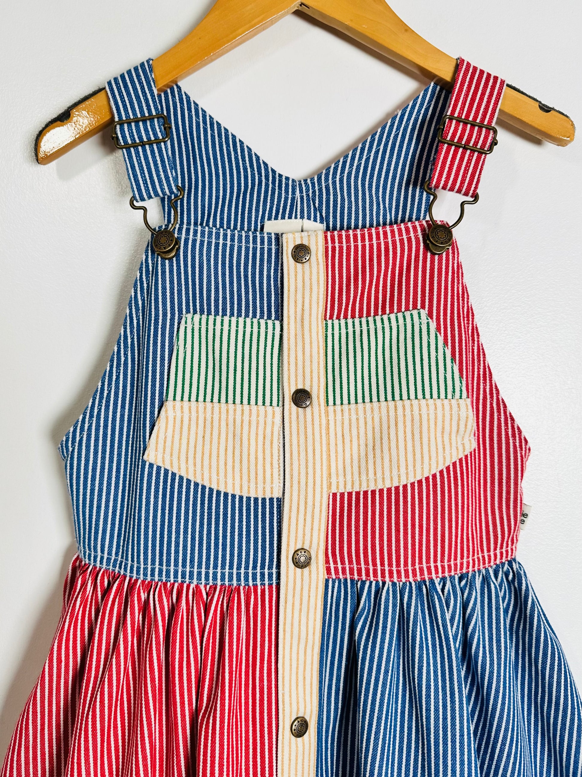 A children's sleeveless dress with patchwork striped fabric panels in blue, red, green, and cream, hanging on a wooden hanger against a plain white wall.