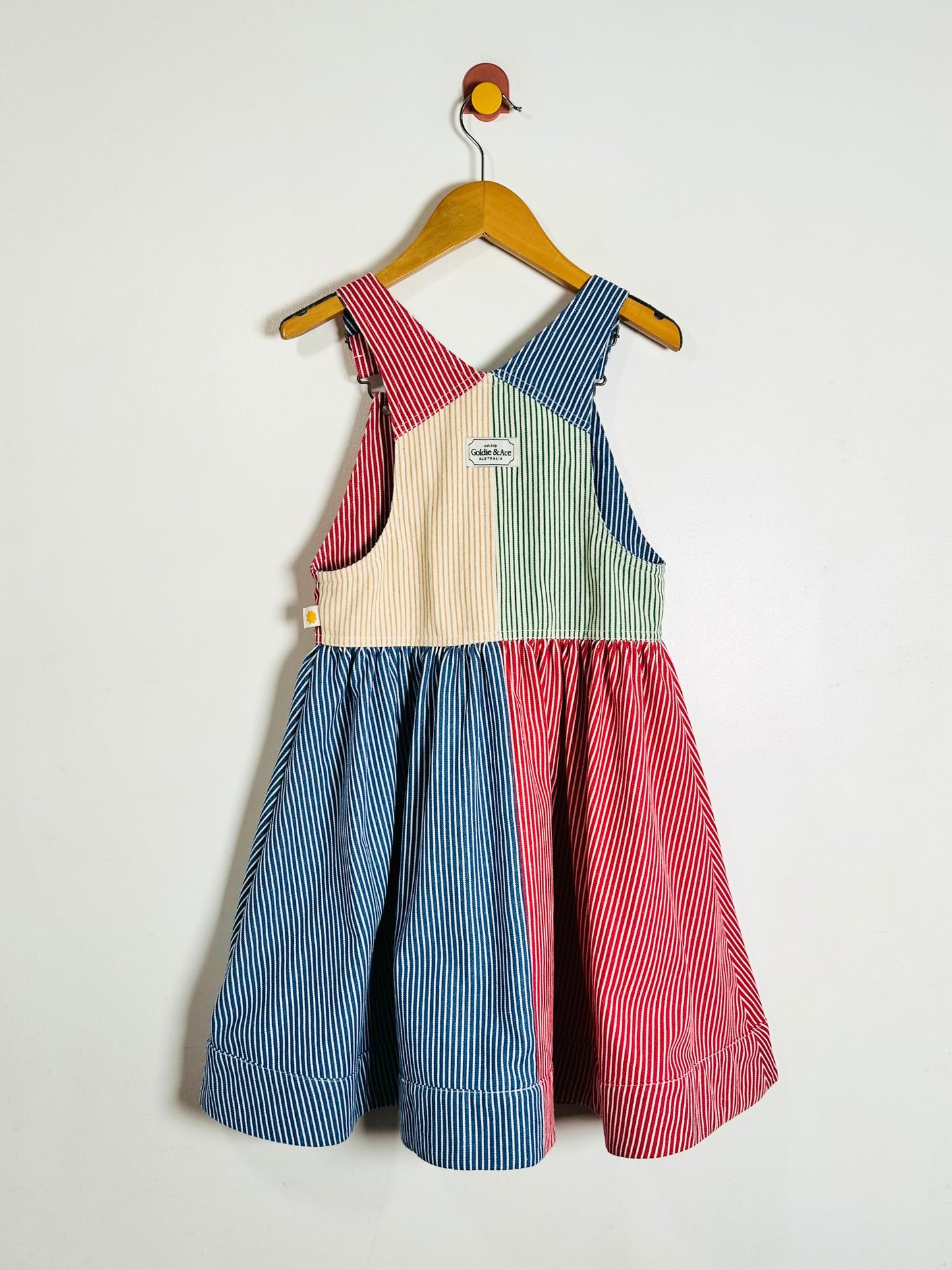 A children's sleeveless pinafore dress with a patchwork design of blue, red, cream, and mint green vertical stripes, hanging on a wooden hanger against a plain white wall.