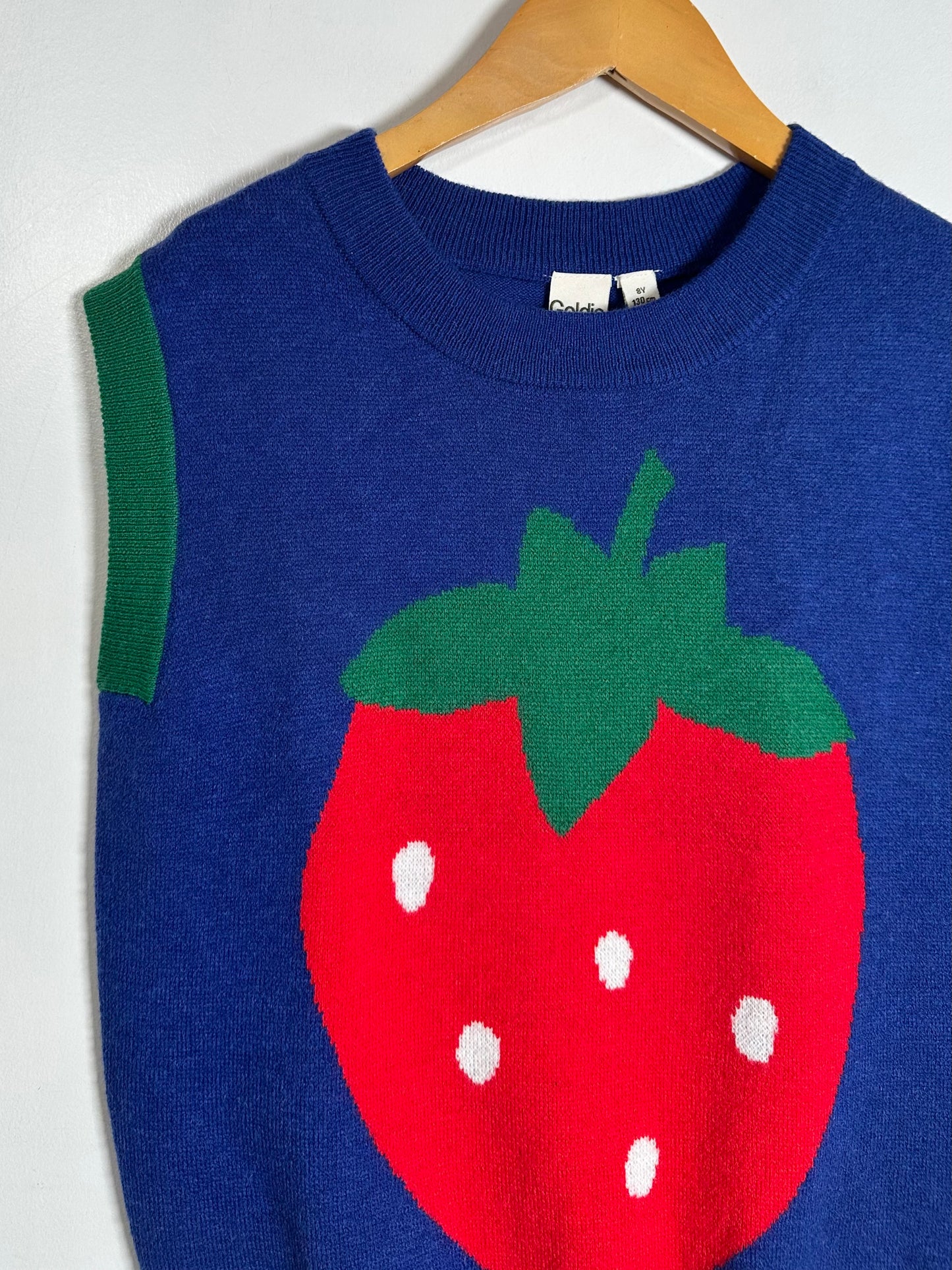 A blue knit sweater vest with a large red strawberry graphic on the front, featuring green leaves and white seeds, hanging on a wooden hanger against a white wall.