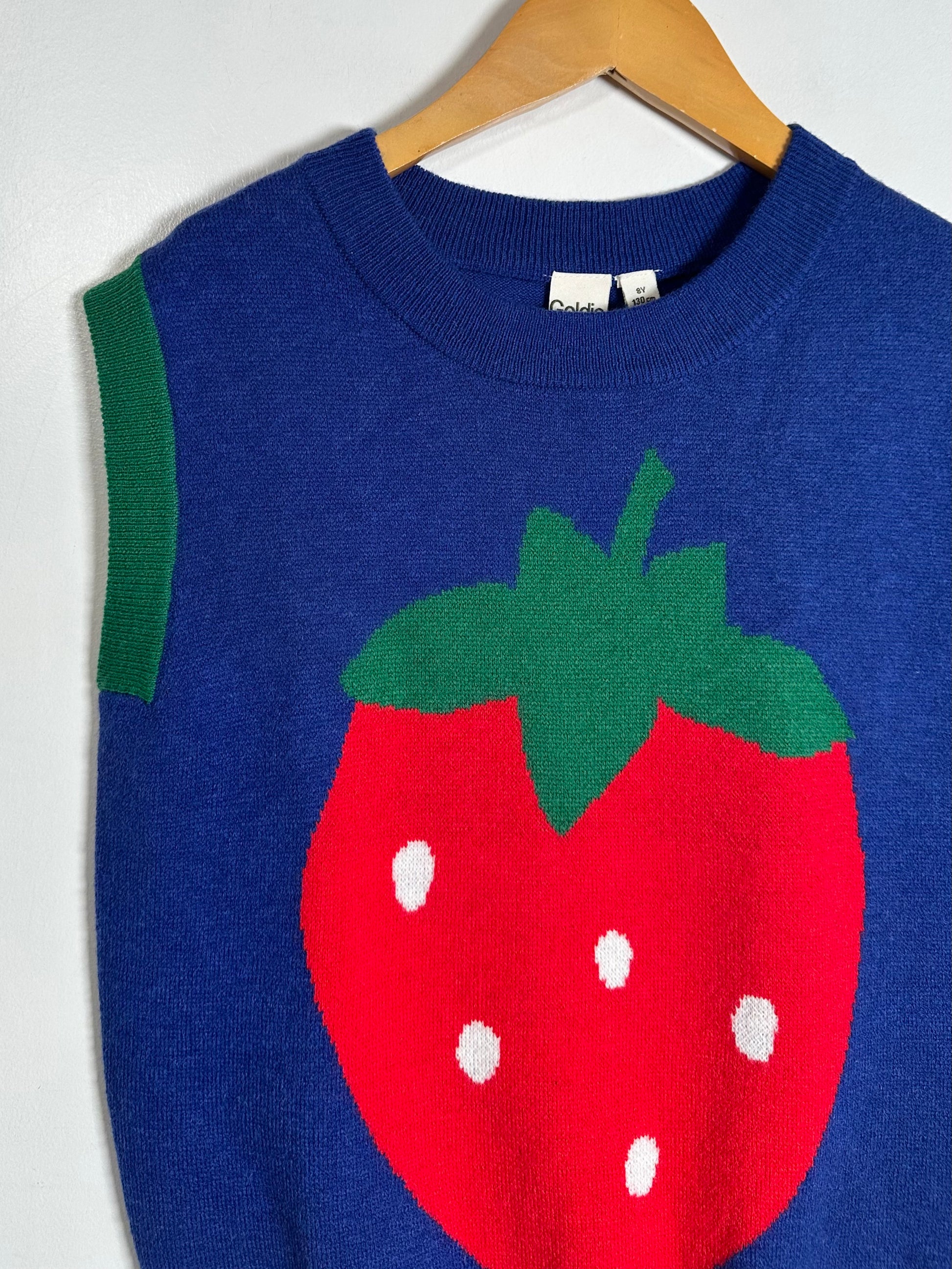A blue knit sweater vest with a large red strawberry graphic on the front, featuring green leaves and white seeds, hanging on a wooden hanger against a white wall.