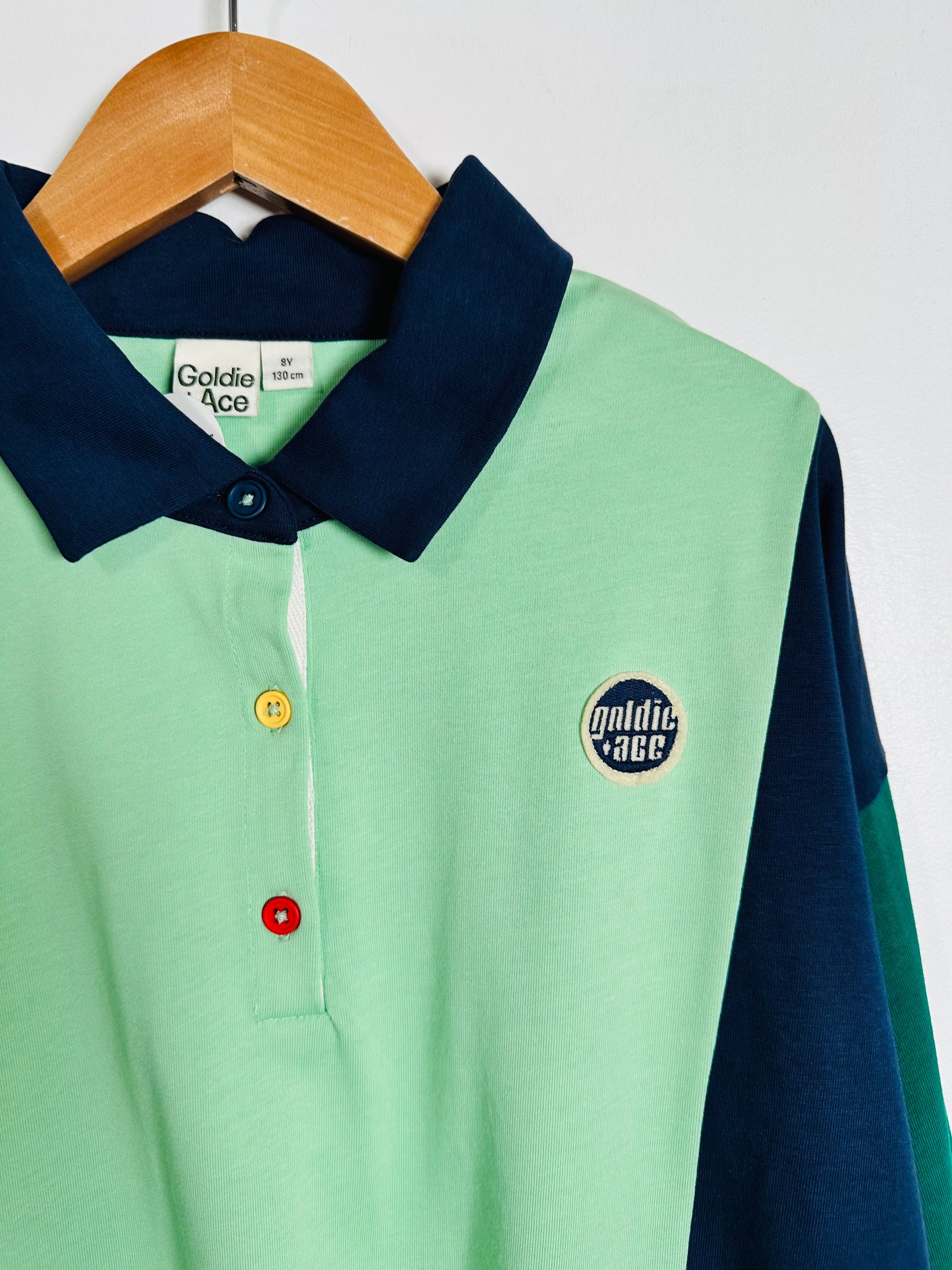 A color-blocked long-sleeve polo shirt by Goldie Ace hanging on a wooden hanger against a white wall. The shirt features a mint green body, a dark navy blue collar, and navy blue sleeves, with colorful buttons (yellow, red) and a circular embroidered logo on the chest.