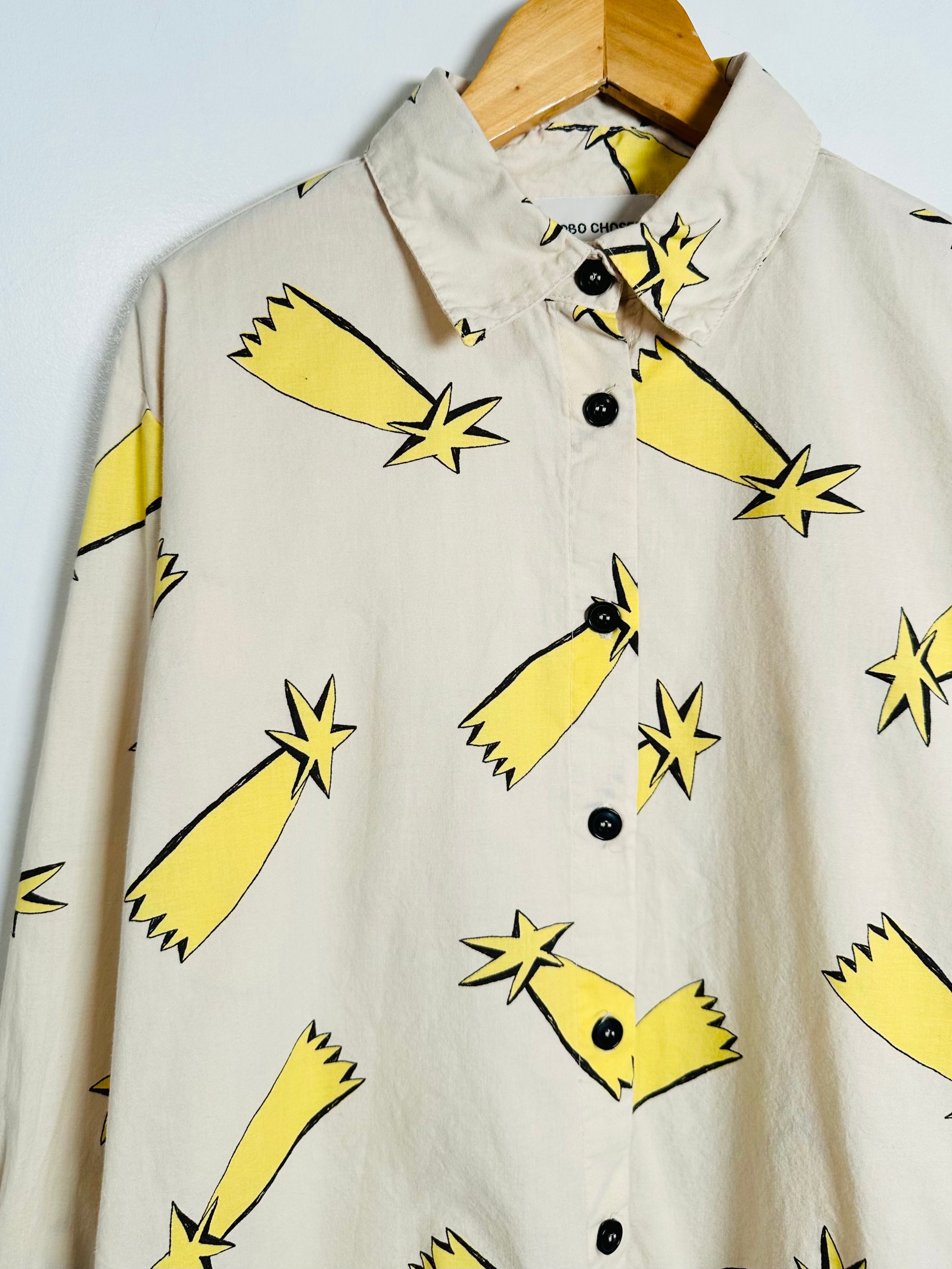 A beige long-sleeve button-up shirt with a yellow and black rocket and star pattern, hanging on a wooden hanger against a light gray background.