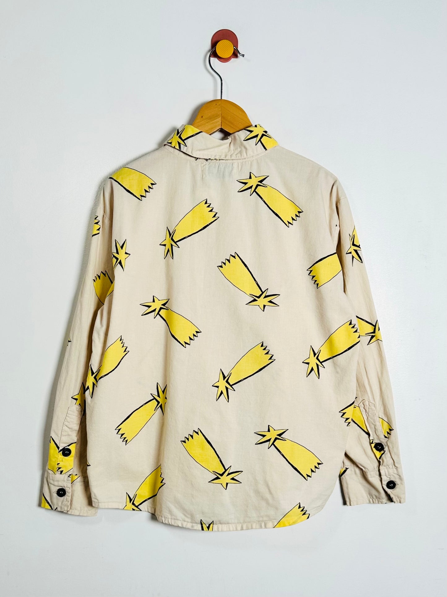 A beige long-sleeve button-up shirt with a yellow and black star print, hanging on a wooden hanger against a plain white wall.
