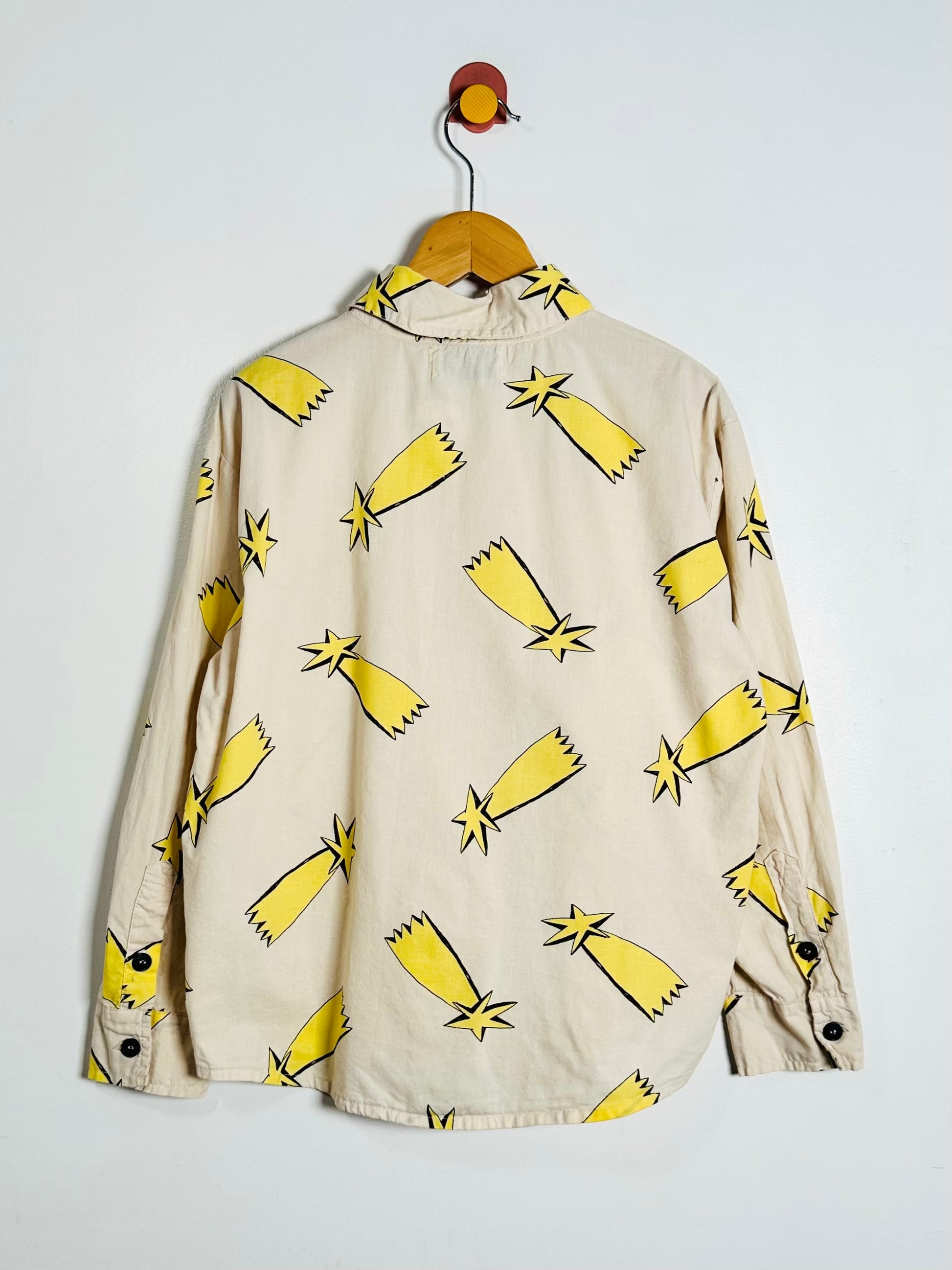 A beige long-sleeve button-up shirt with a yellow and black star print, hanging on a wooden hanger against a plain white wall.