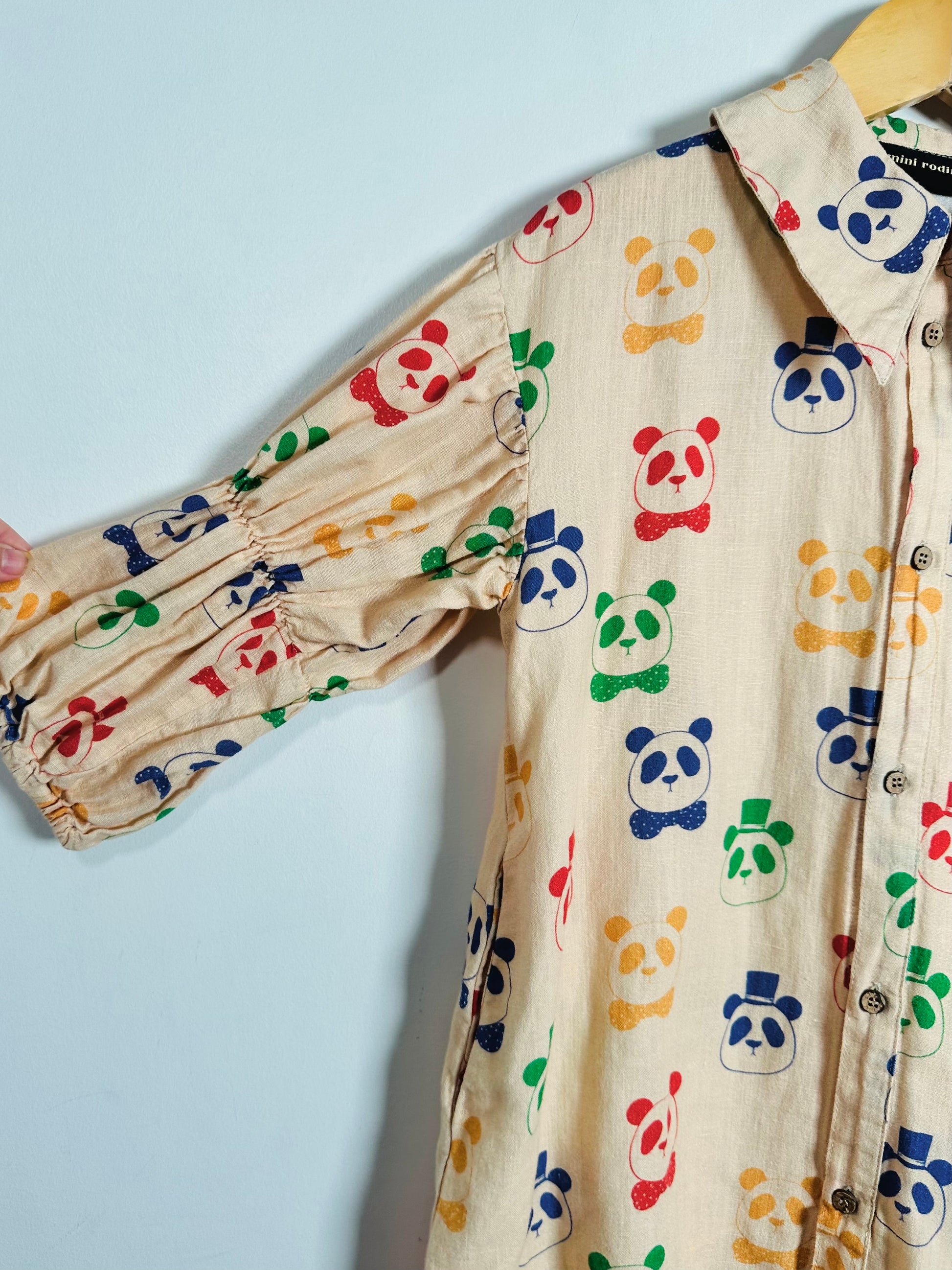 A cream-colored cotton dress featuring a colorful print of cartoon panda faces in red, blue, green, and yellow. The shirt has a pointed collar, button-down front, and gathered elbow-length sleeves, shown hanging on a wooden hanger against a light blue background.