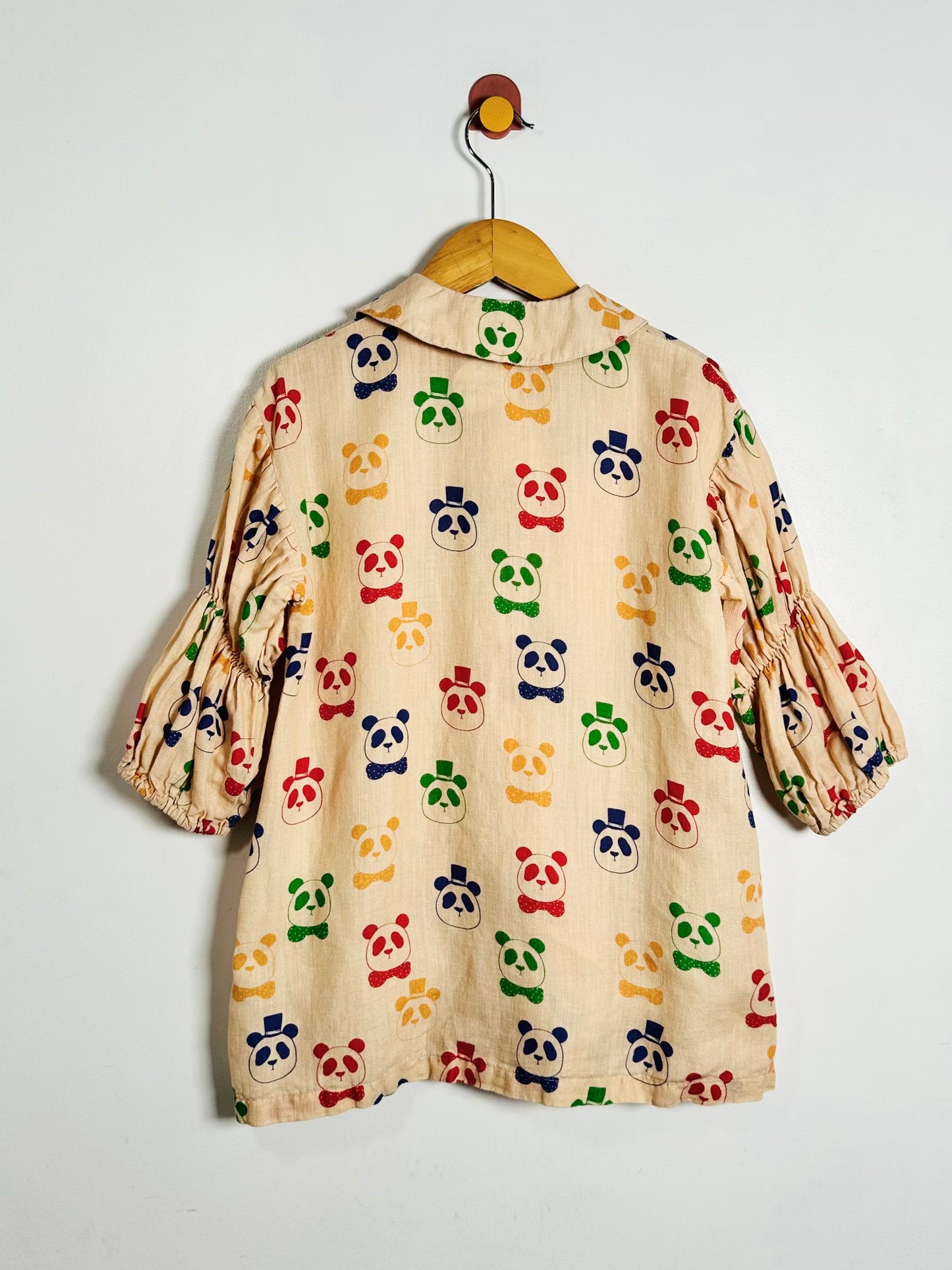 A beige cotton dress with a colorful panda print, hanging on a wooden hanger against a plain white wall.
